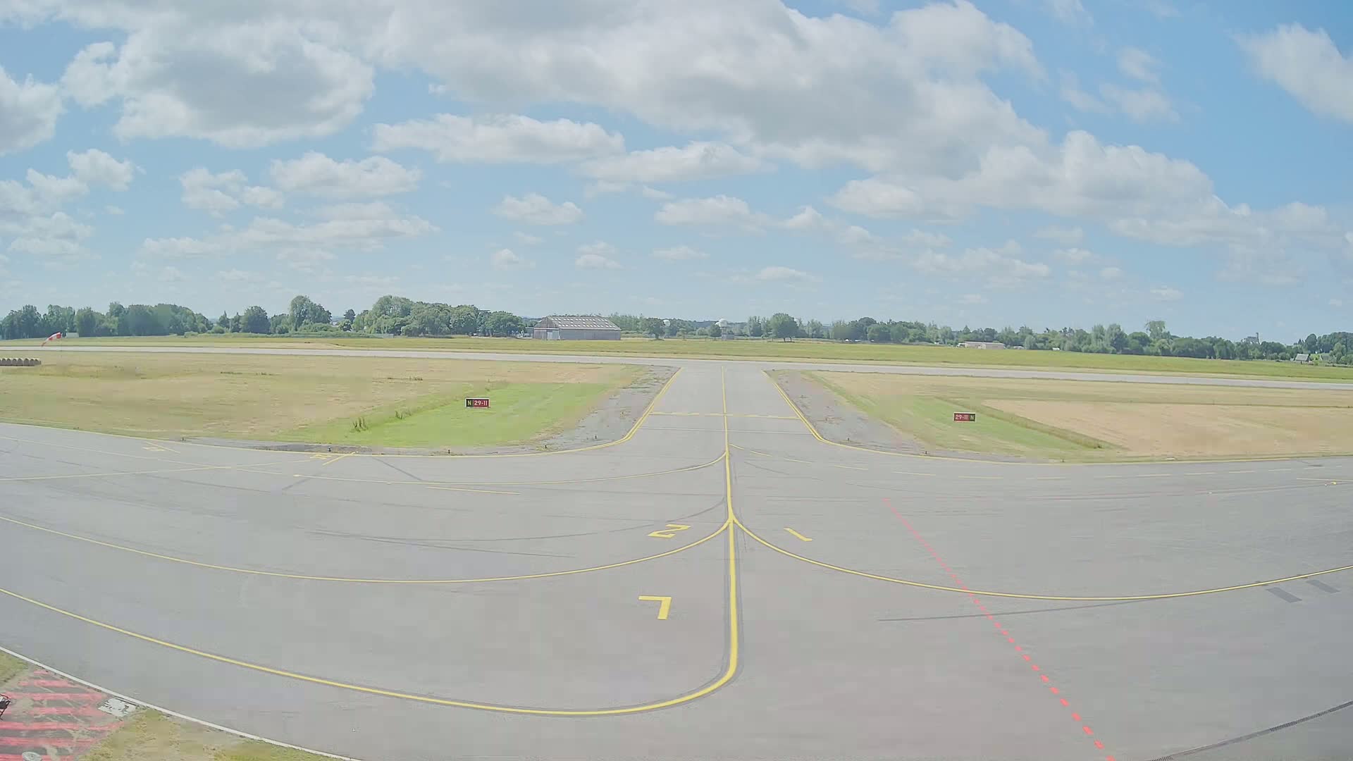 A tarmac runway at a small airport on a partly cloudy day with a grassy field and trees in the background.