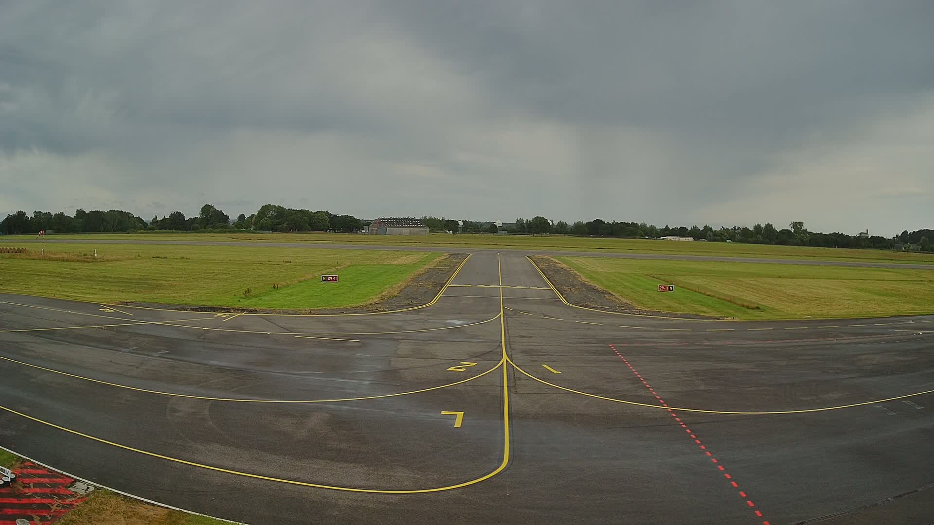 A tarmac airfield with yellow lines marking taxiways and runways, surrounded by grassy fields under an overcast sky.