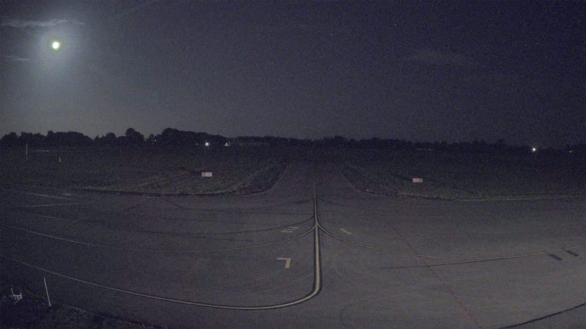 Under a bright moonlit night sky, a dark tarmac runway stretches into a field bordered by a treeline.