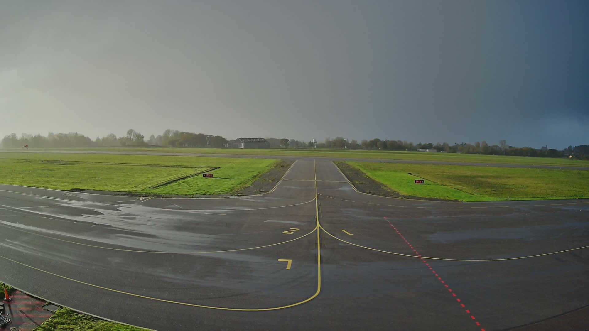 A wet airport tarmac, marked with yellow lines, is flanked by green grassy fields leading to a distant tree line and buildings, all under a cloudy sky that darkens from light grey on the left to a stormy grey on the right.
