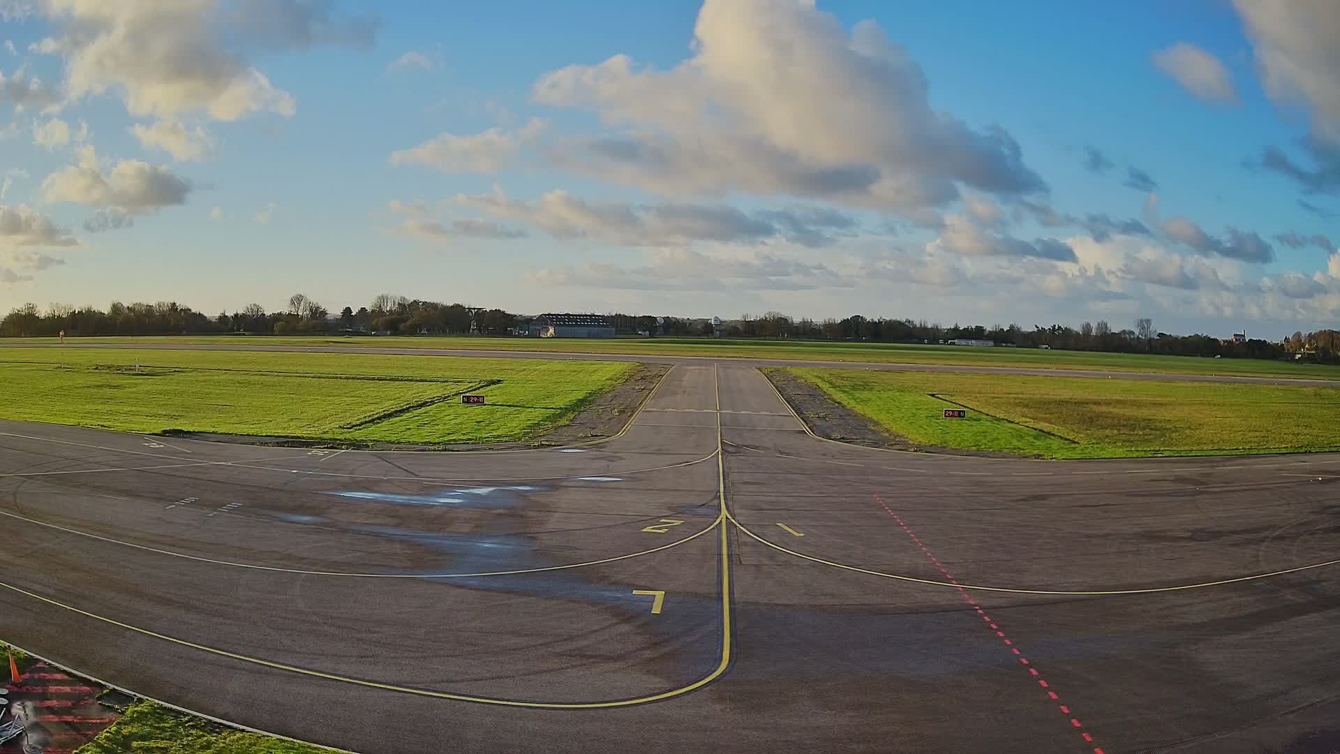 An empty airport taxiway with yellow and red markings, bordered by vibrant green grass, extends towards a distant runway under a bright, partly cloudy blue sky.