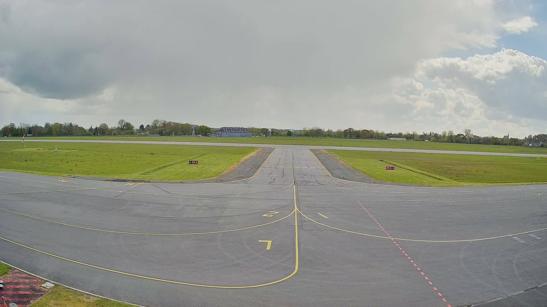 An empty airport taxiway with yellow and red markings, bordered by vibrant green grass, extends towards a distant runway under a bright, partly cloudy blue sky.