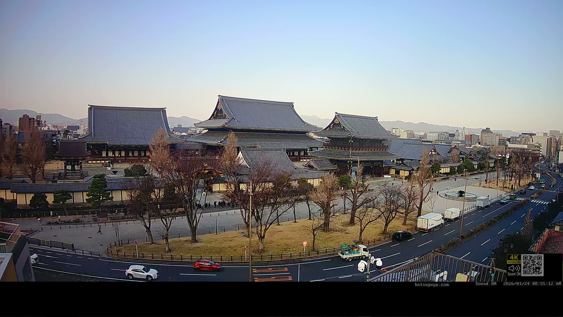 An elevated view reveals a sprawling complex of traditional Japanese temple buildings with dark tiered roofs, surrounded by bare trees, roads with light traffic, and a distant city skyline backed by mountains, all under a clear and calm sky.