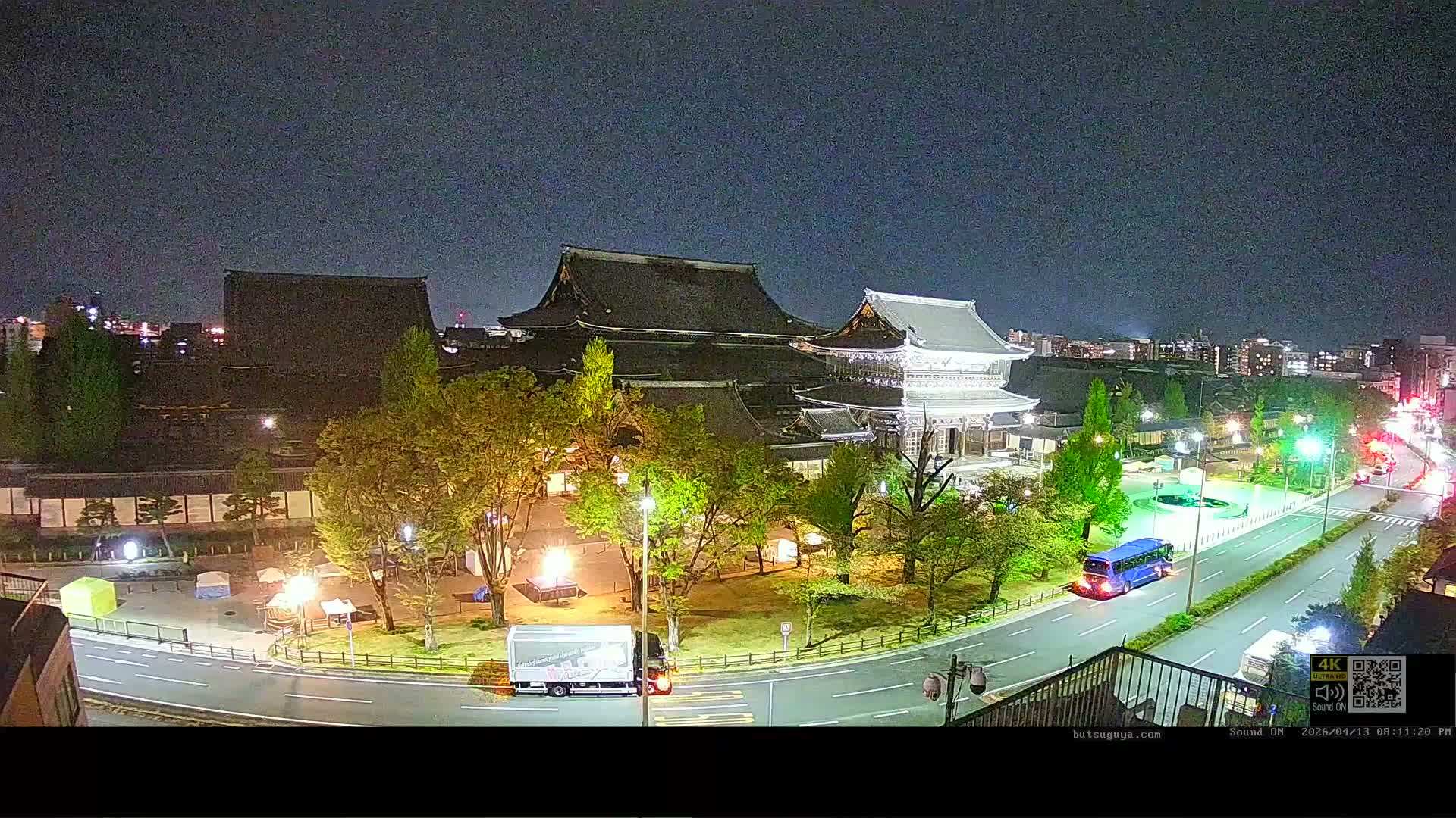 An elevated view reveals a sprawling complex of traditional Japanese temple buildings with dark tiered roofs, surrounded by bare trees, roads with light traffic, and a distant city skyline backed by mountains, all under a clear and calm sky.