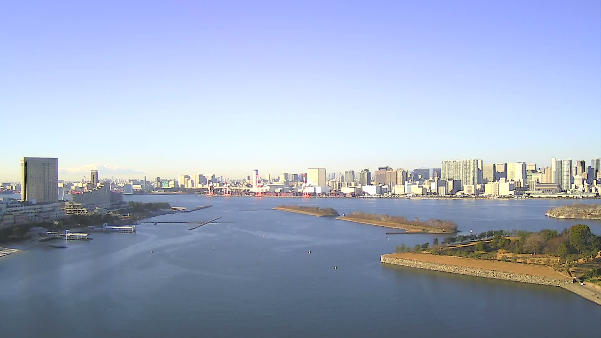 A panoramic view shows a wide bay with calm blue water, dotted with small islands and buoys, leading to a sprawling city skyline featuring numerous buildings and a distant snow-capped mountain, all under a clear, sunny blue sky.