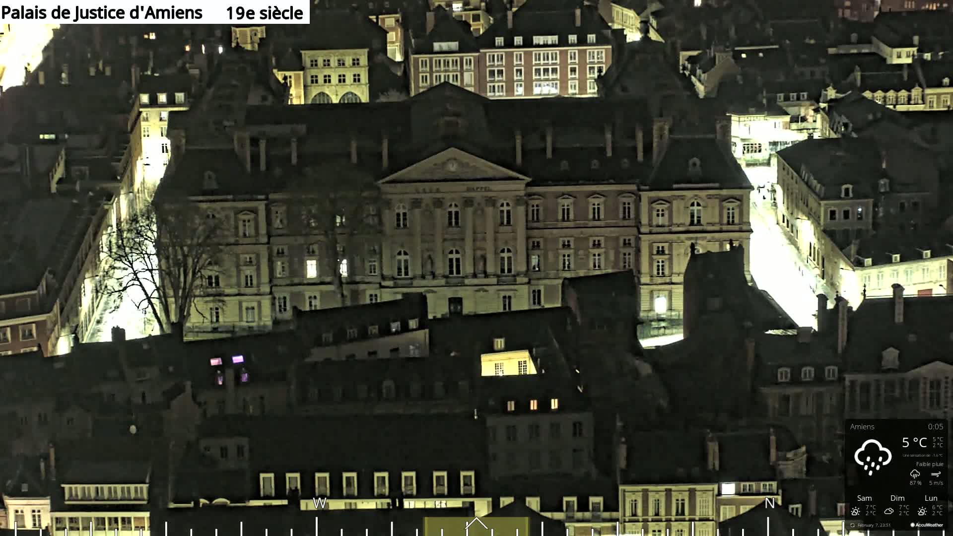 A nighttime aerial view of a European city shows a large, illuminated building surrounded by other smaller buildings, with light rain falling.