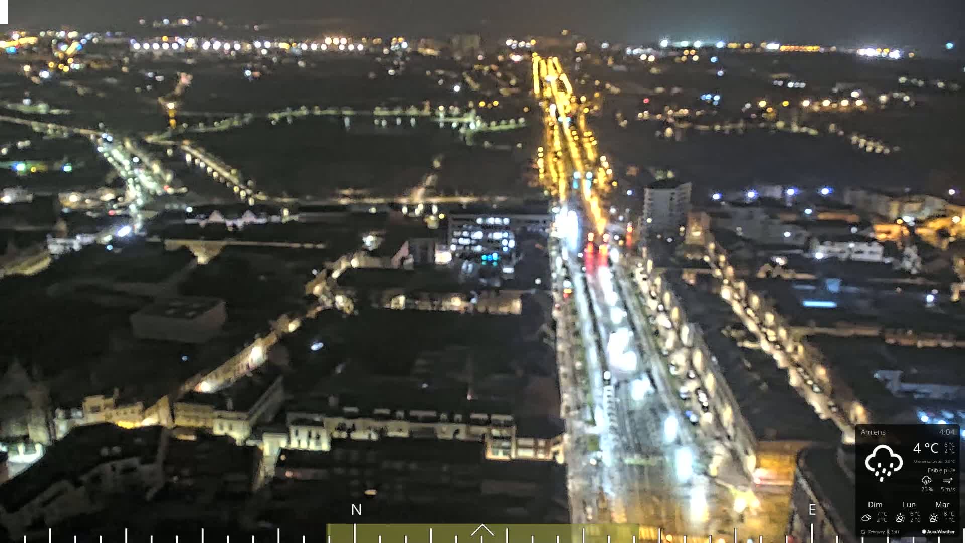 A nighttime aerial view of a city with a brightly lit main road and surrounding streets, under light rain.