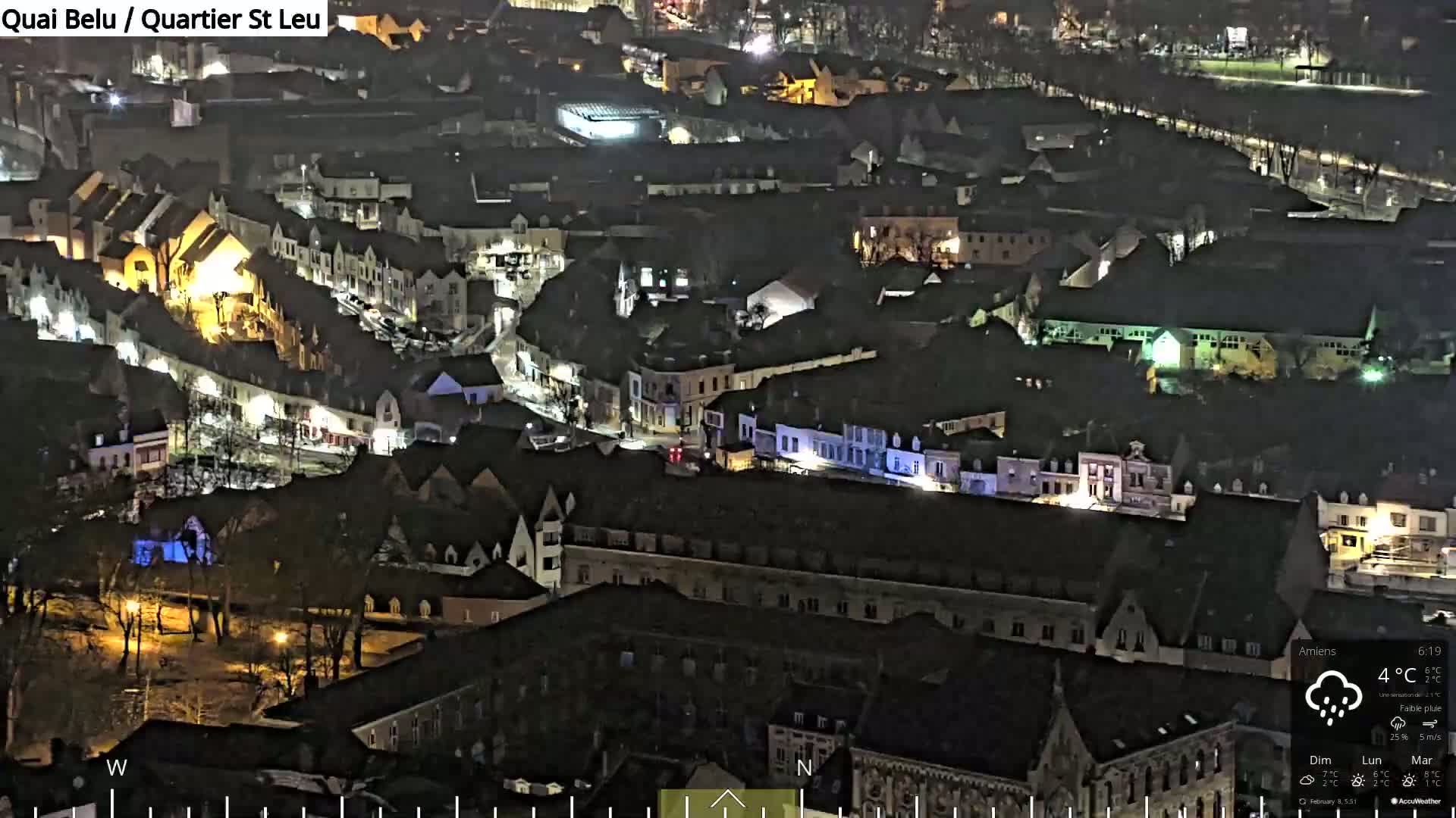 A nighttime, aerial view of a town with light rain, showing many buildings illuminated by streetlights and residential lights.