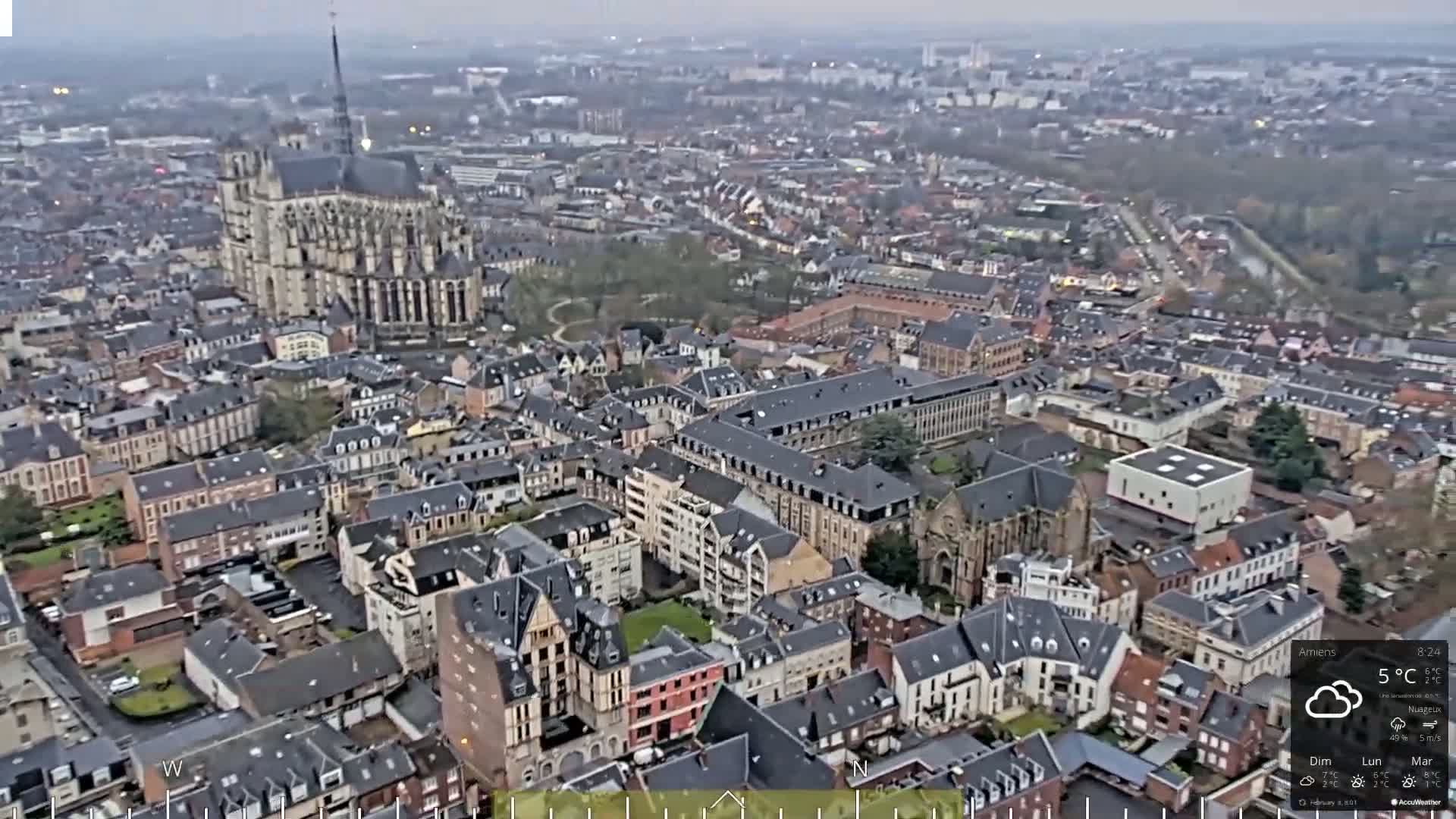 An aerial view shows a European city on an overcast day, with a large cathedral prominently featured in the center.