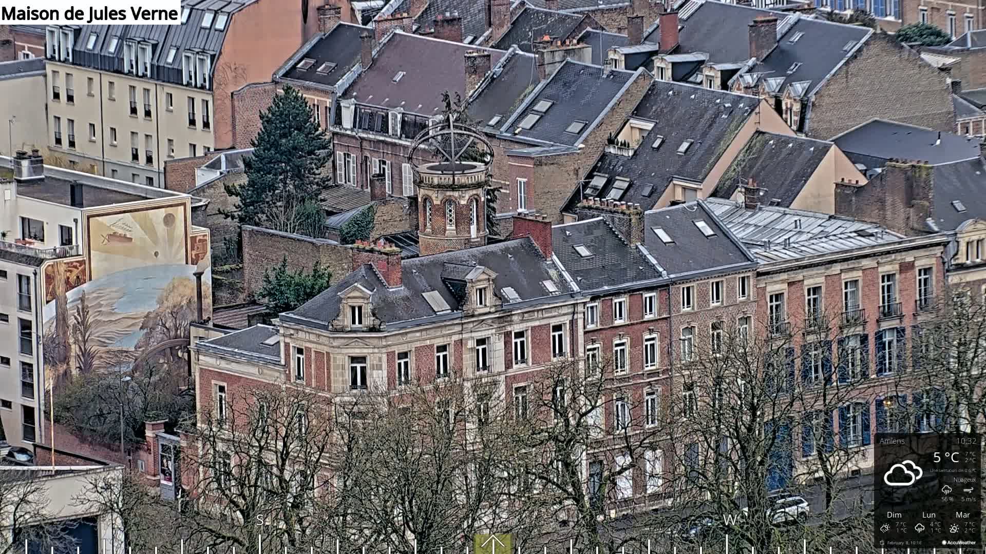 A high-angle view of a European city on a cloudy day shows a dense collection of buildings with dark gray slate roofs, punctuated by a multi-story brick building with a unique tower and a large mural depicting a ship and balloon.