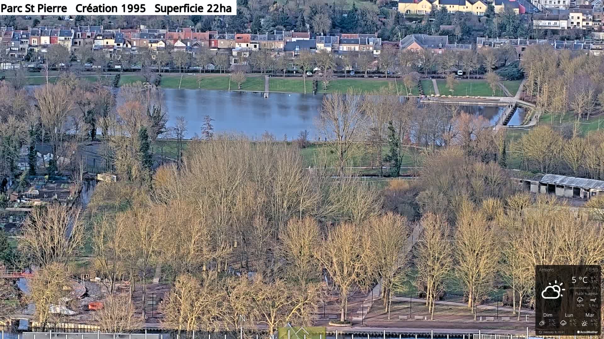 A mostly cloudy day reveals a park with a large pond, many bare trees, and residential buildings in the background.