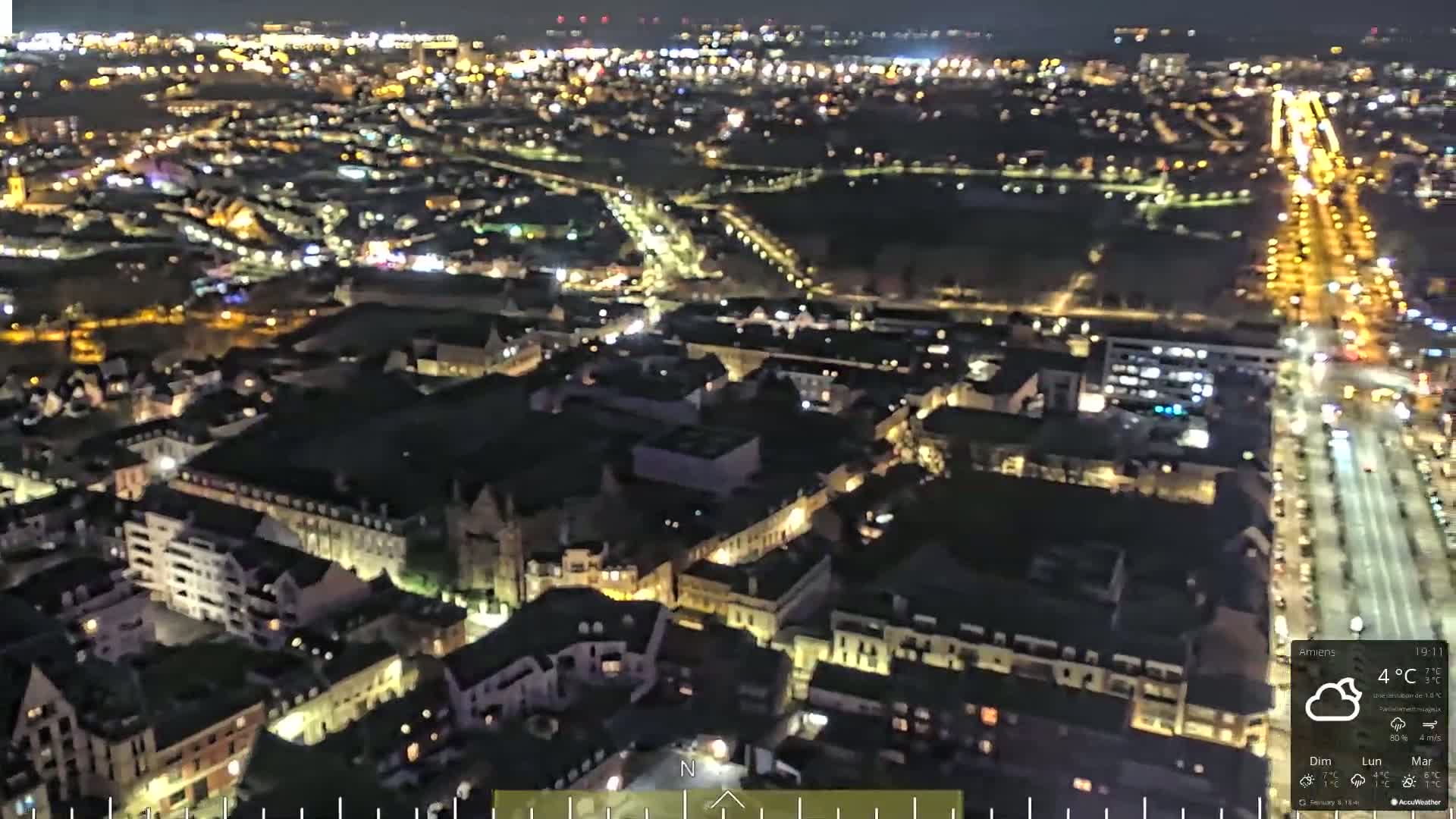 A nighttime aerial view of a city with brightly lit streets and buildings under cloudy skies.
