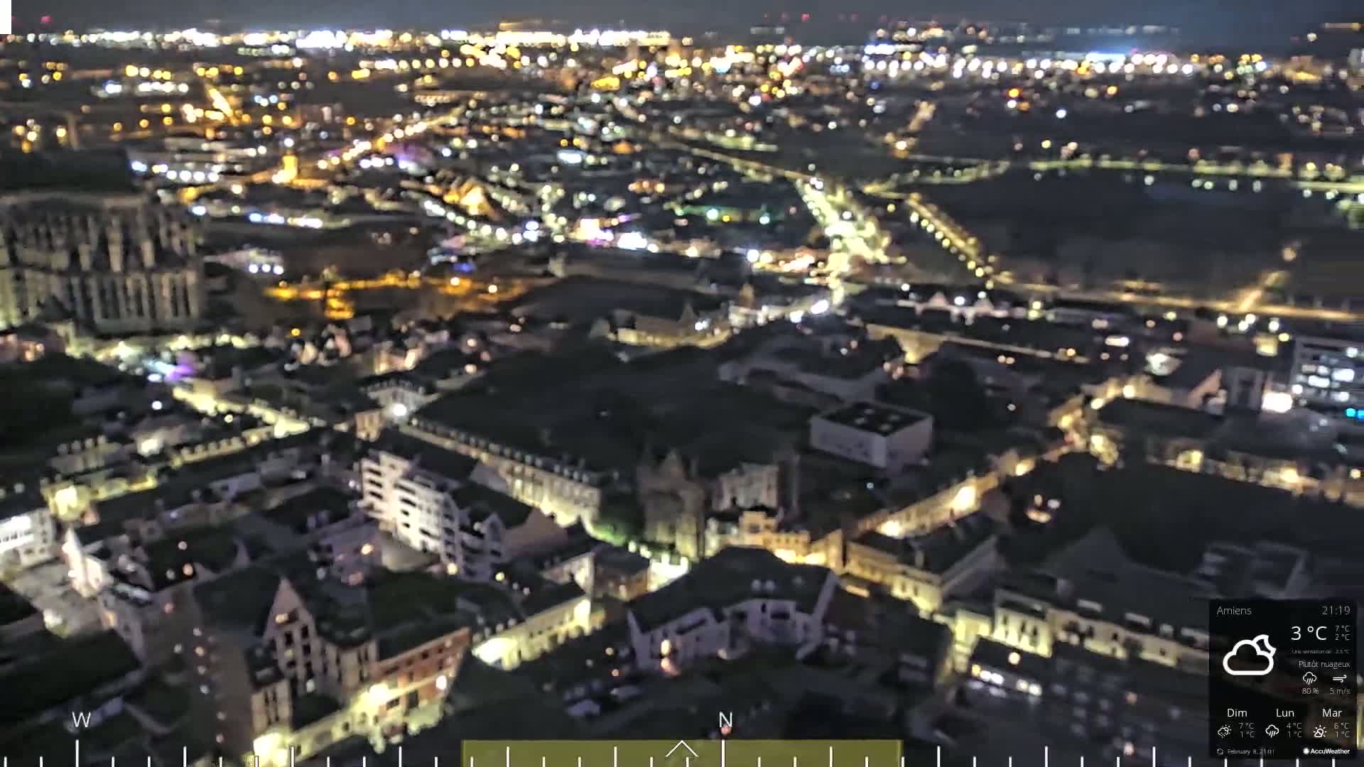 A nighttime aerial view of a city with many illuminated buildings and streets under partly cloudy skies.