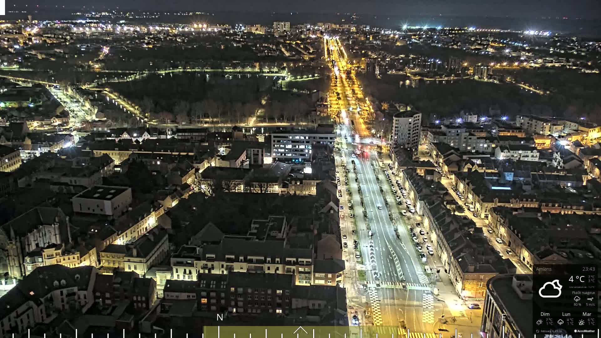 A nighttime aerial view of a city with a long, brightly lit street running through it, under partly cloudy skies.