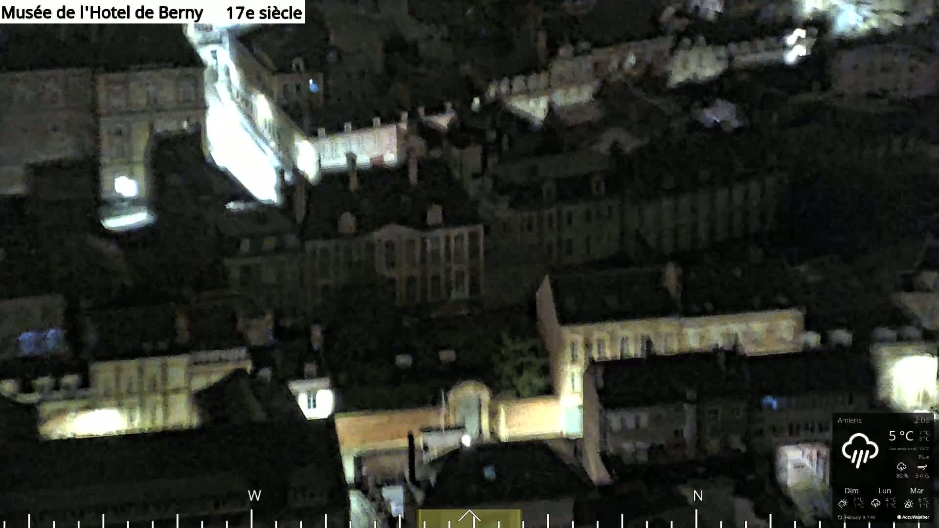 A nighttime aerial view of a city with dimly lit buildings under a cloudy sky with rain.