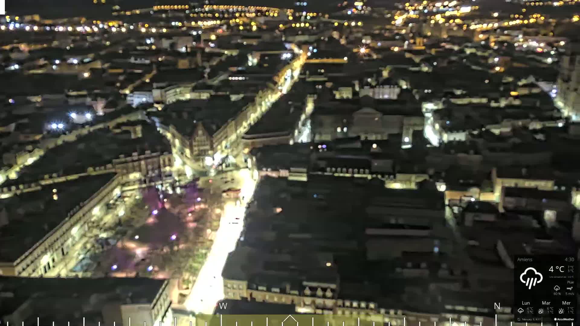 An aerial nighttime view of a city with illuminated streets and buildings under a cloudy sky with rain.