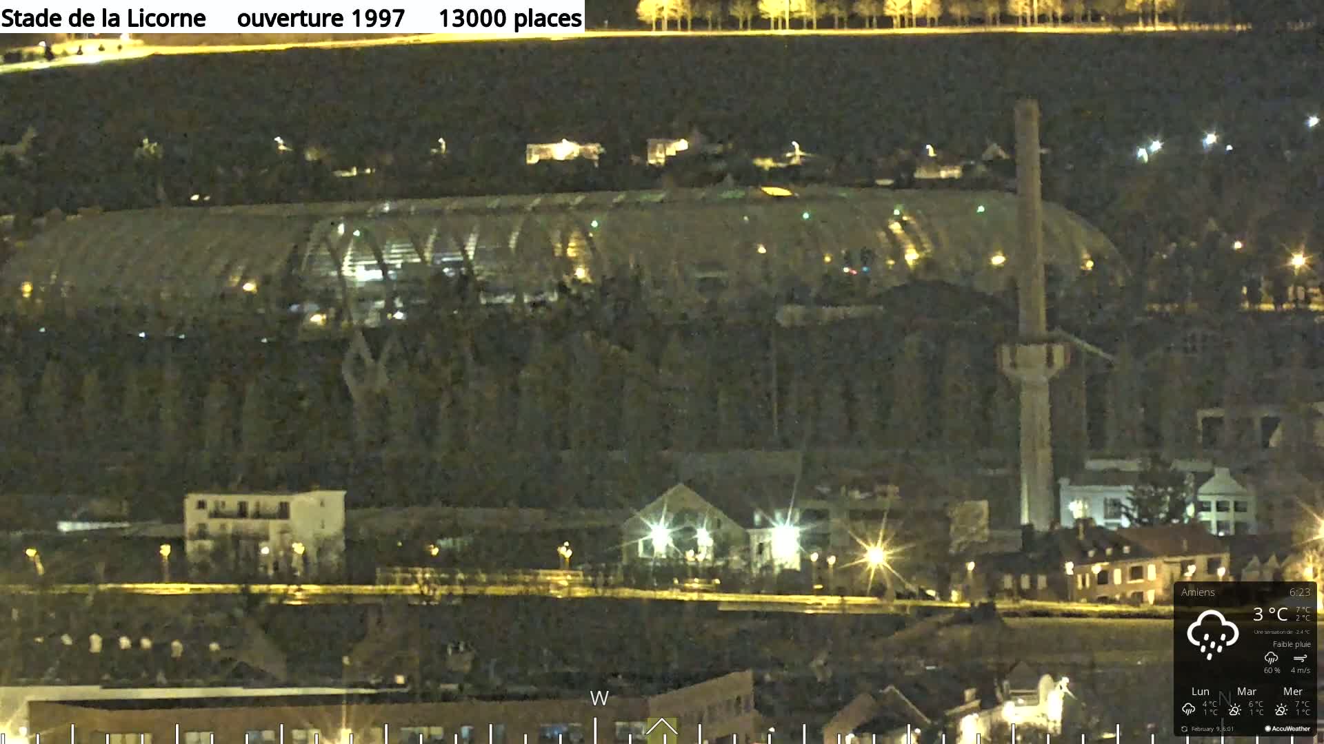 A nighttime view of a large stadium with lights on, surrounded by a city with streetlights, under a slightly overcast sky with light rain.