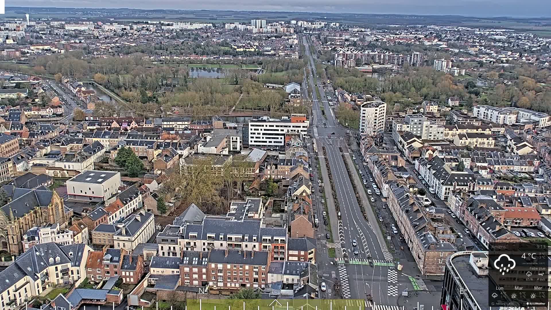 A high-angle, long shot of a city on an overcast day, showing a long road lined with parked cars and buildings on either side, extending to a wider cityscape in the distance.