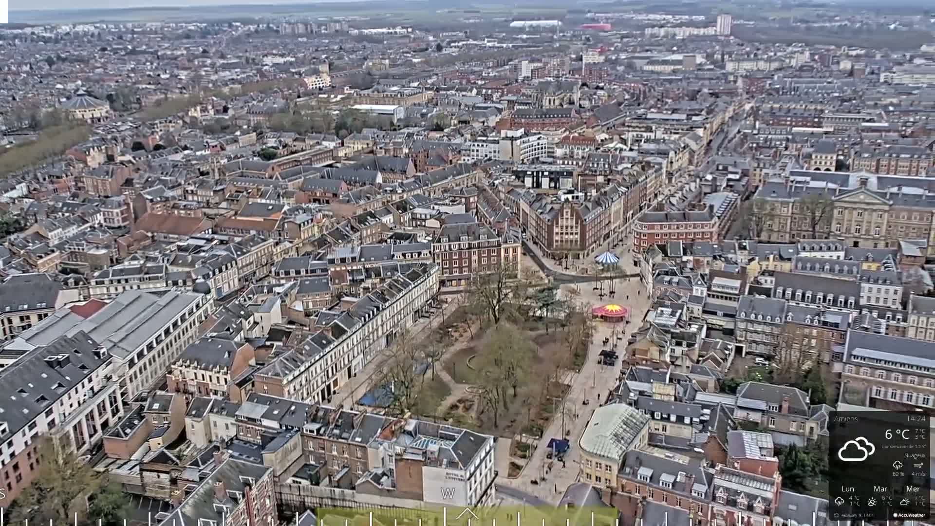 An aerial view of a European city on an overcast day shows a large town square with a small carousel and a park surrounded by closely-packed buildings.