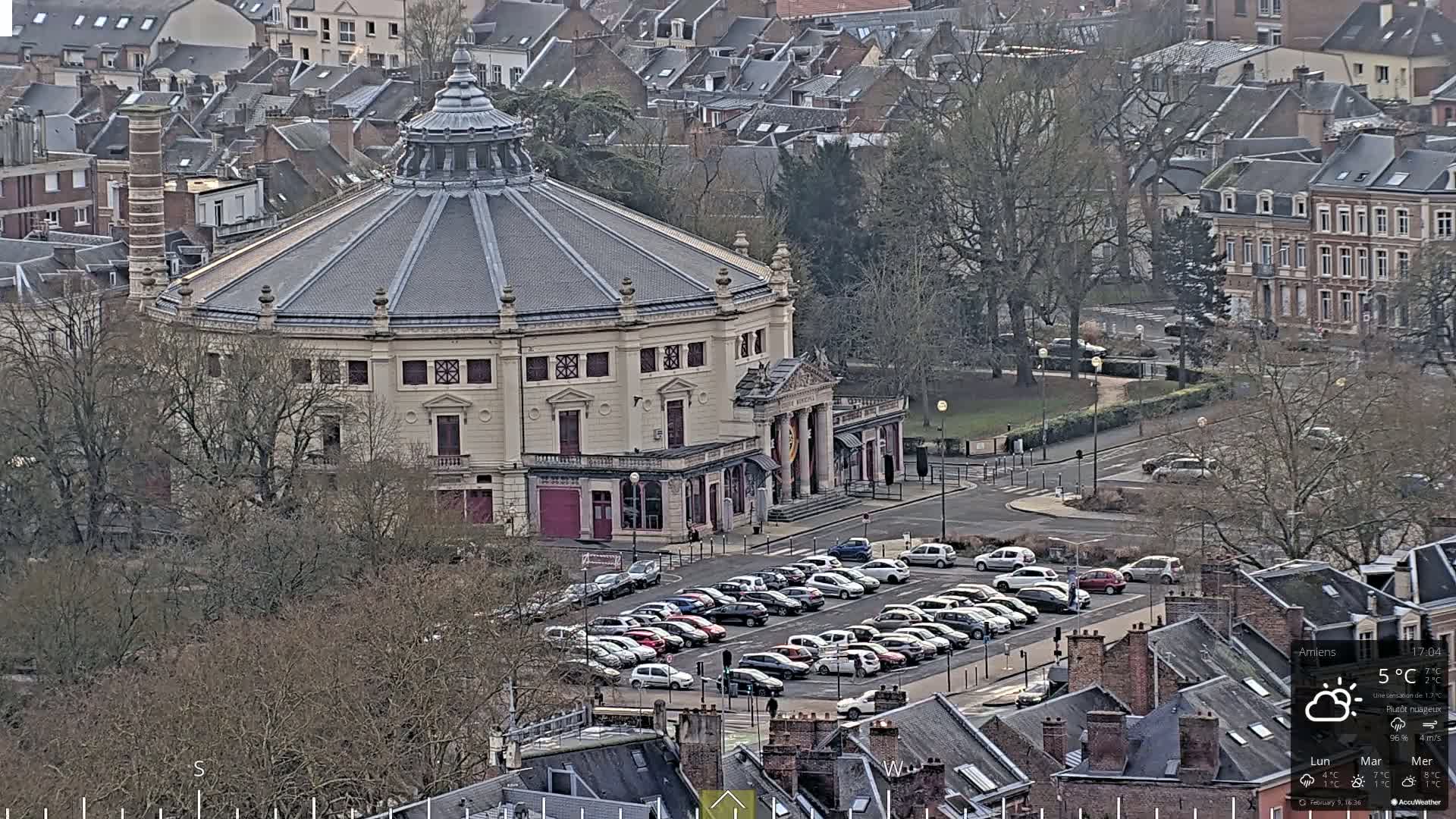 A large, round, light-colored building with a dark dome roof sits in a city parking lot on a mostly cloudy day.