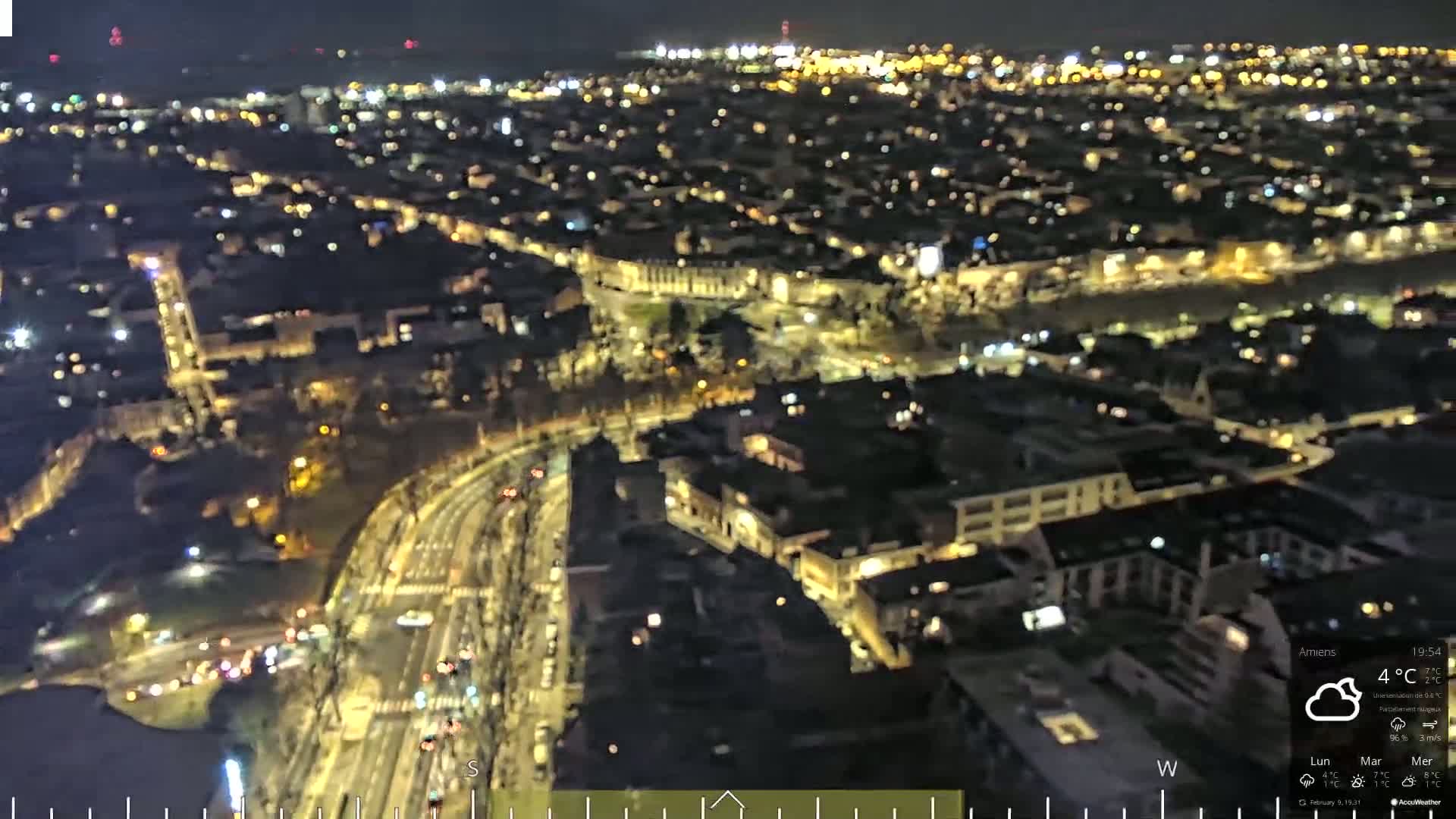 A nighttime, aerial view of a city with dimly lit buildings and roadways showing vehicle traffic under cloudy conditions.