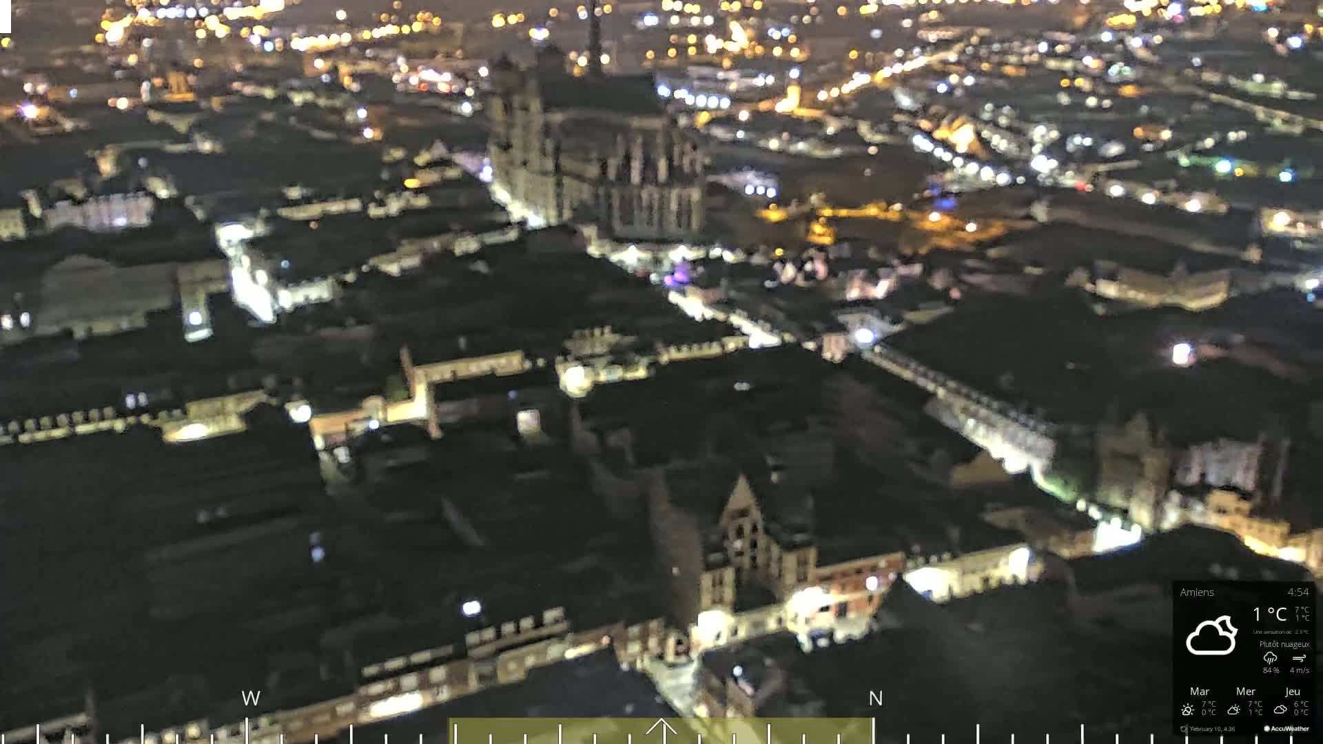 A nighttime aerial view of a city with numerous illuminated buildings and streets, under partly cloudy skies.
