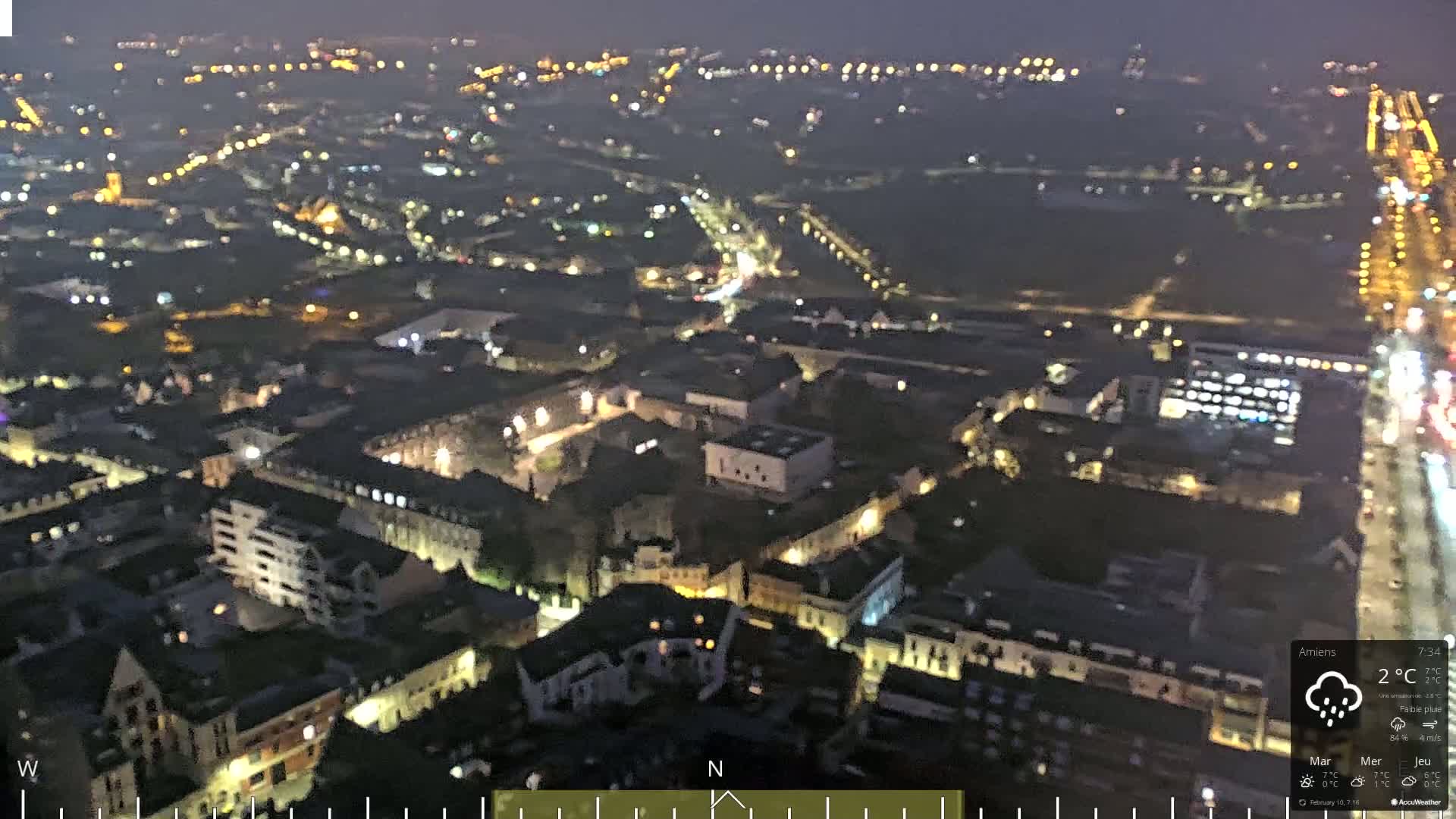 A nighttime, high-angle view of a city with light rain, showing numerous illuminated buildings and streets.