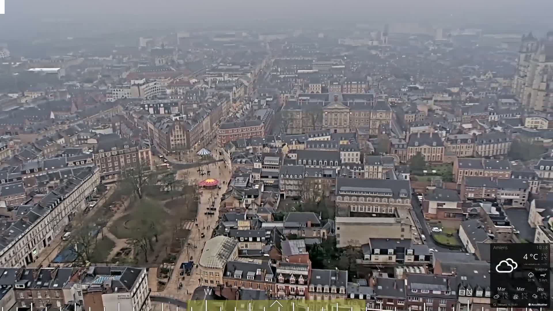 A high-angle, long shot of a European city on an overcast day shows many buildings of varying heights and styles, a central square with a small carousel, and some green spaces.