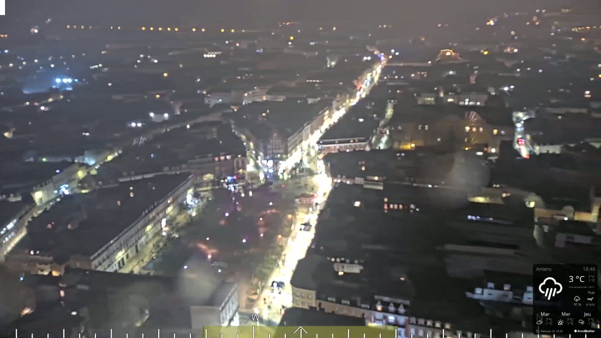 A nighttime aerial view of a city street illuminated with traffic and building lights under rainy conditions.