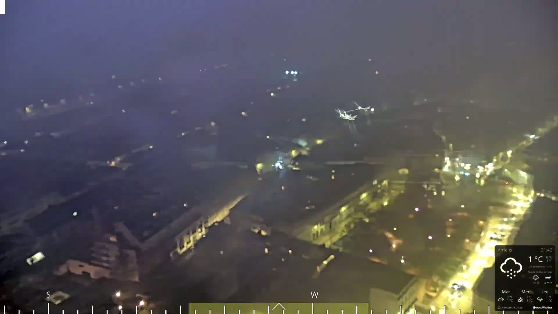 A nighttime aerial view of a city illuminated by streetlights under a hazy, snowy sky with lightning visible in the distance.