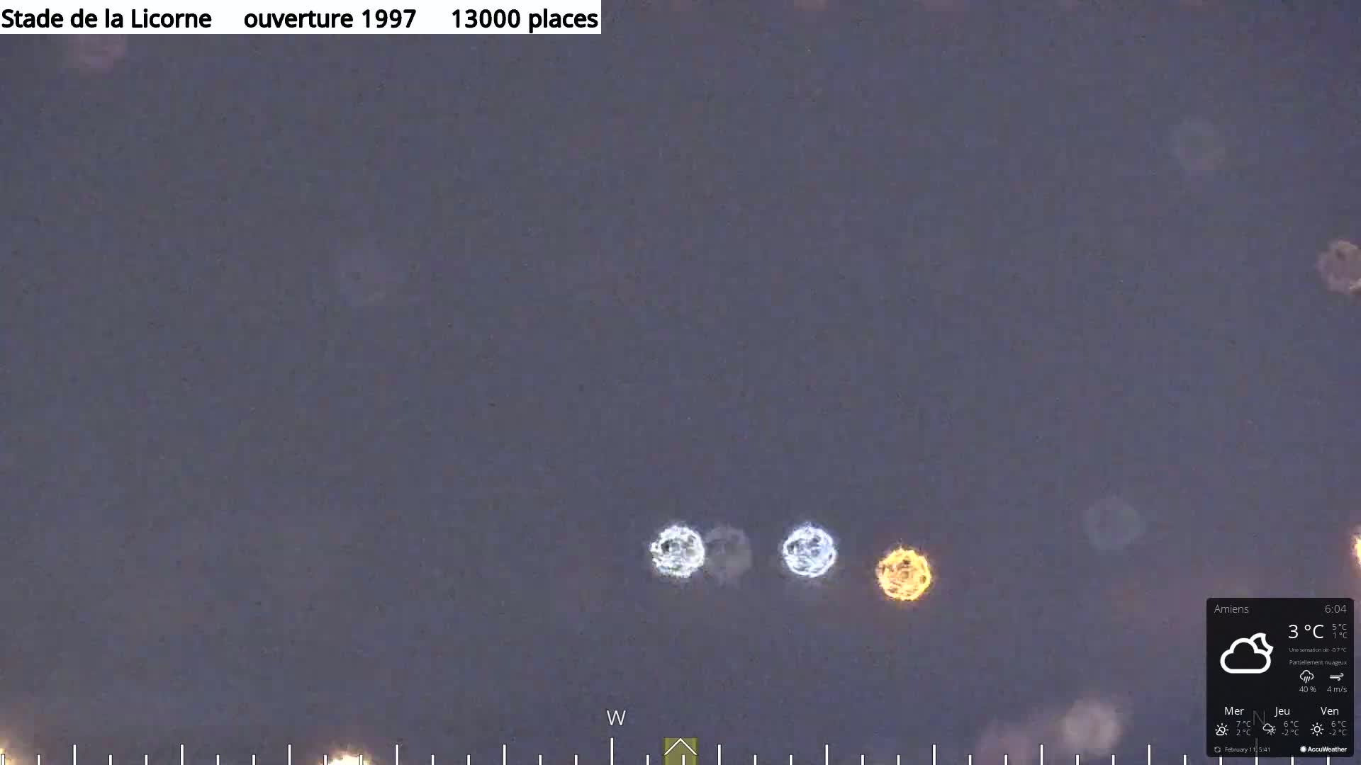 A night-time, partly cloudy view shows three bright, blurry lights of varying colors against a dark sky.