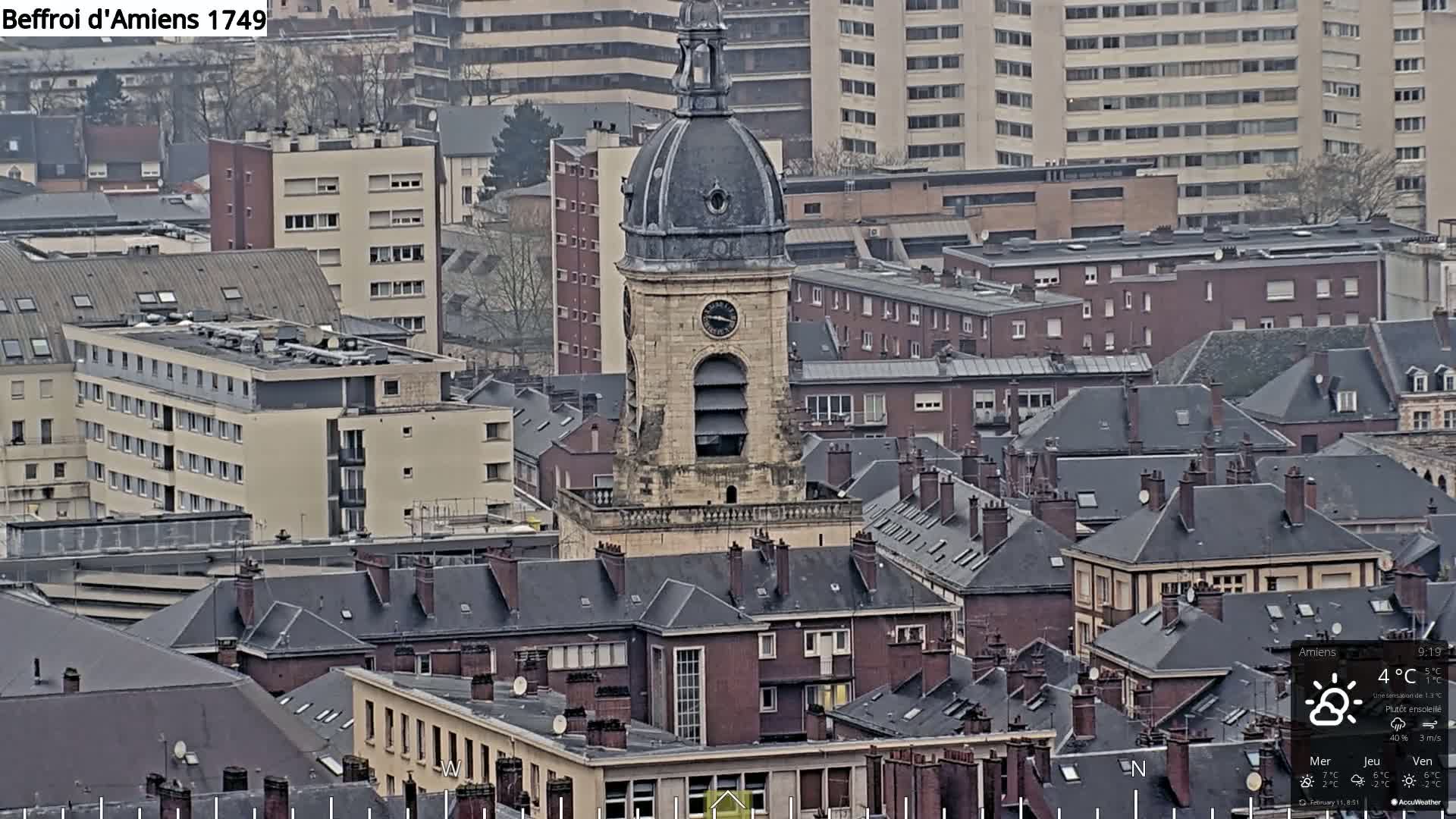 A high-angle, long shot of a city on an overcast day, centered on a tall, domed clock tower amongst many buildings with dark gray rooftops.