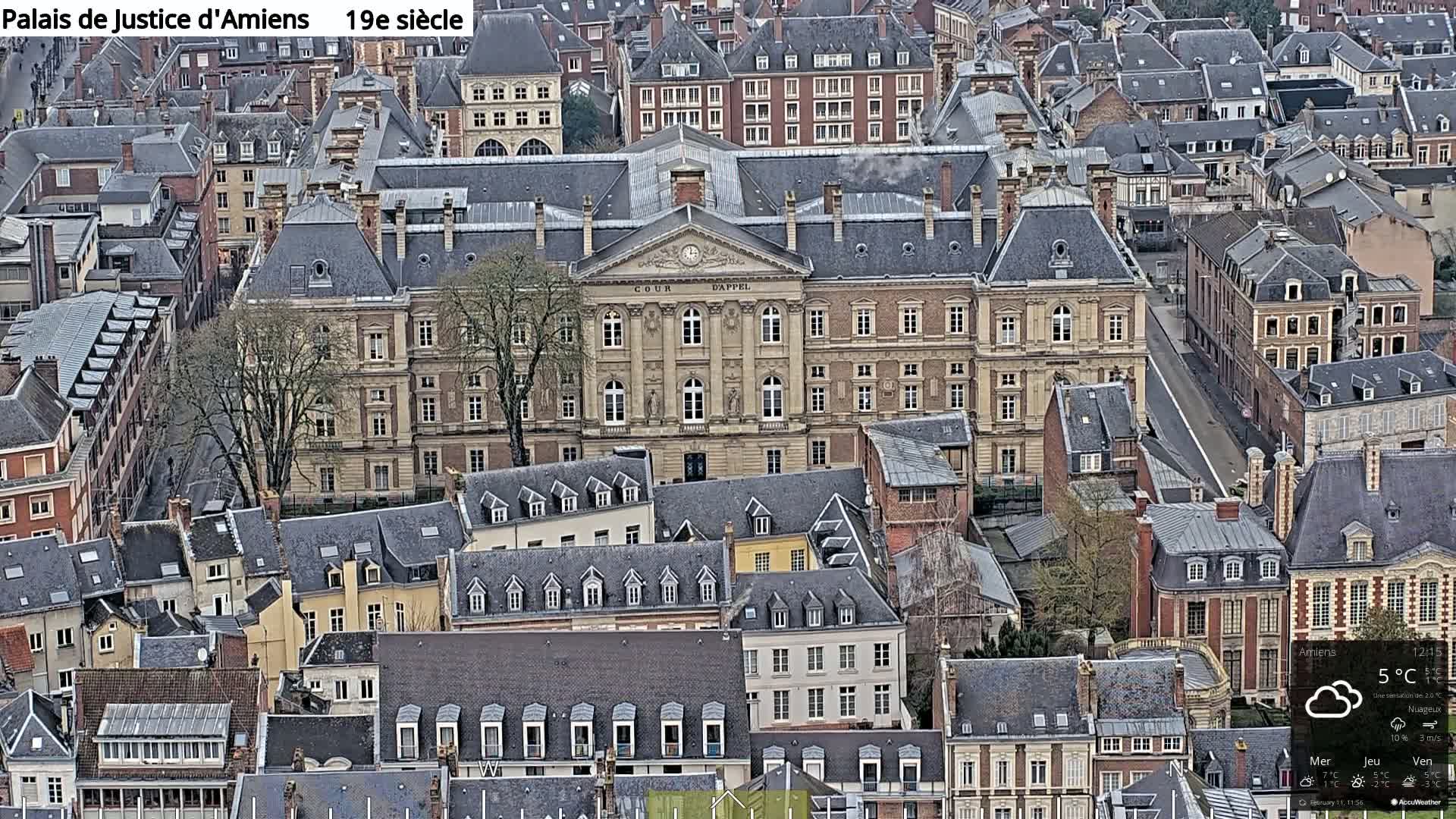 An aerial view of a large, light-brown stone building surrounded by many smaller, dark-roofed buildings on a cloudy day.