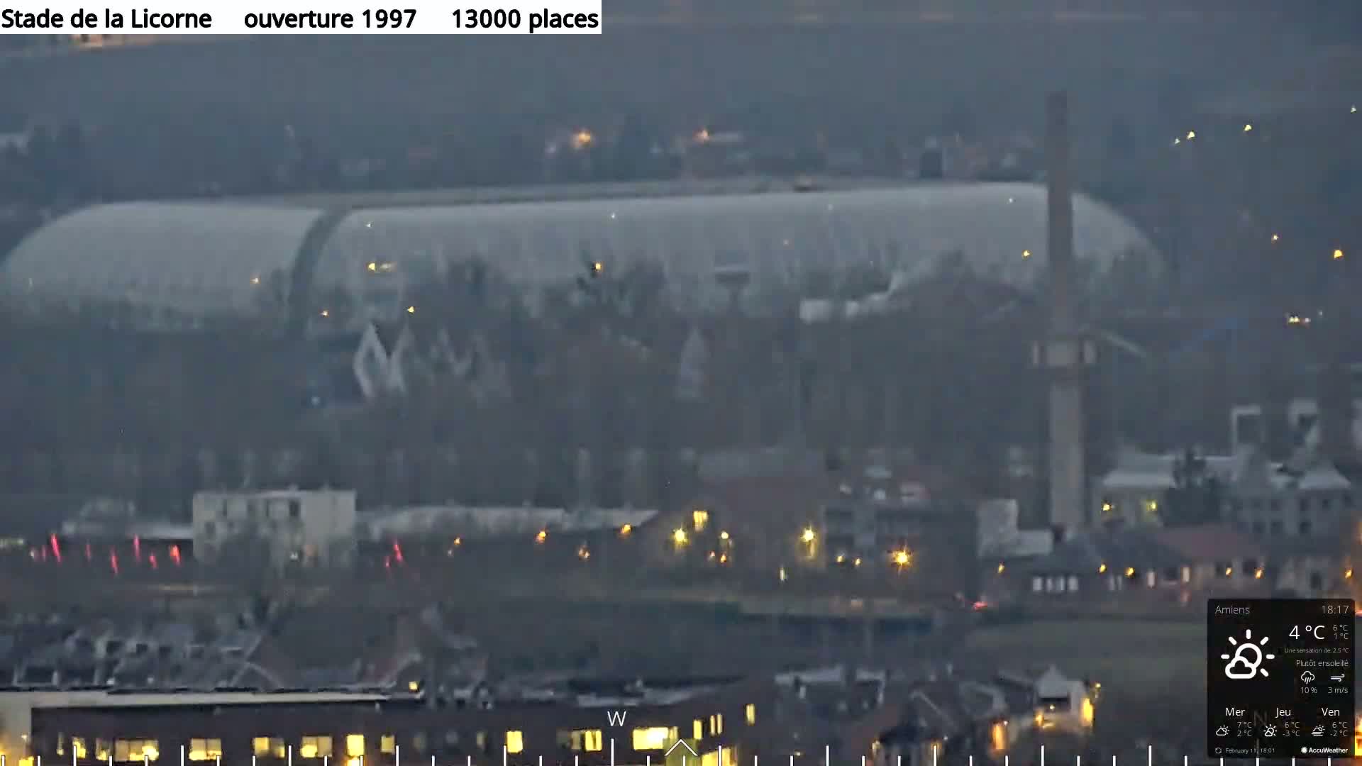 A large stadium with a curved roof is visible in the distance under a mostly clear, dusky sky, with scattered lights illuminating the surrounding town.