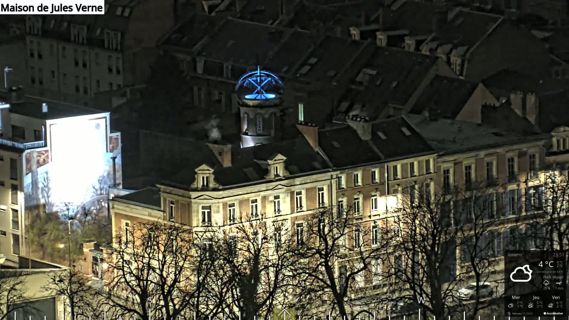 A nighttime aerial view of a city block with a large building illuminated, its rooftop featuring a blue-lit structure, under a mostly clear sky.