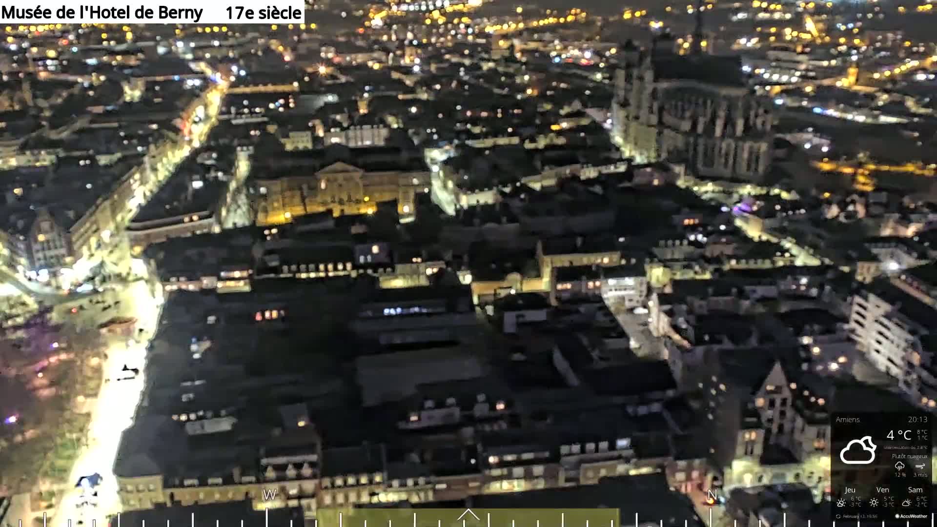 A nighttime aerial view of a city with illuminated streets and buildings, under partly cloudy skies.