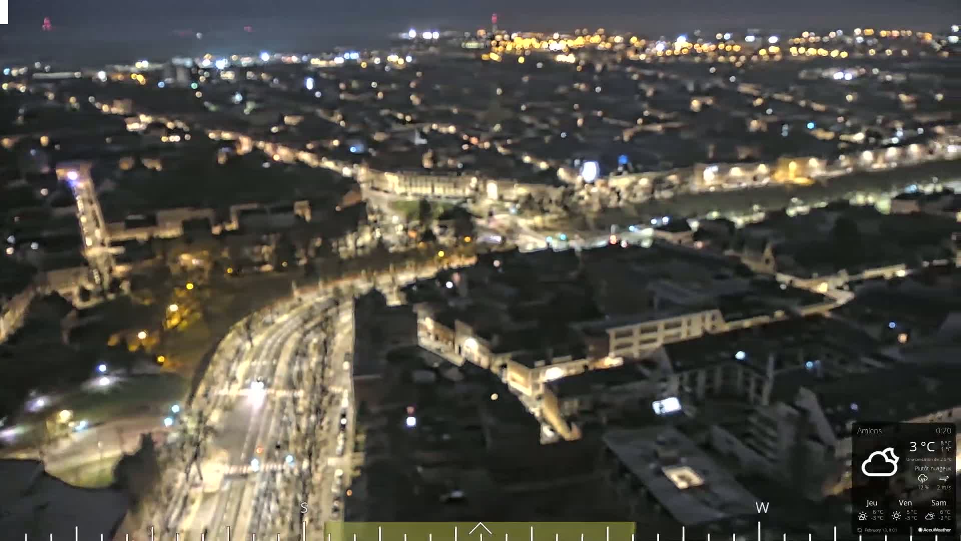 A nighttime, high-angle view of a city with many illuminated buildings and streets, under partly cloudy skies.