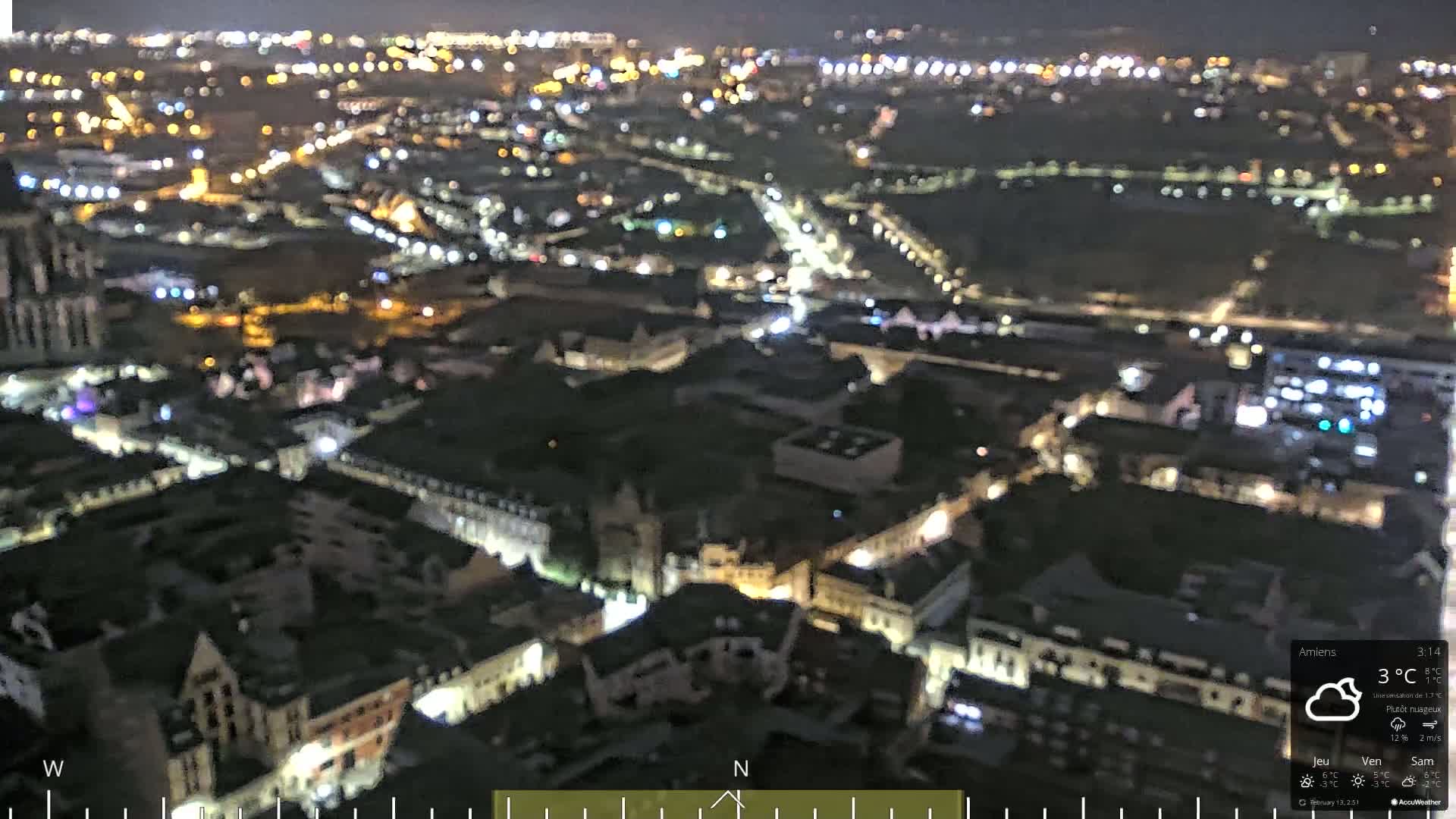 A blurry, nighttime aerial view of a city with illuminated streets and buildings under partly cloudy skies.