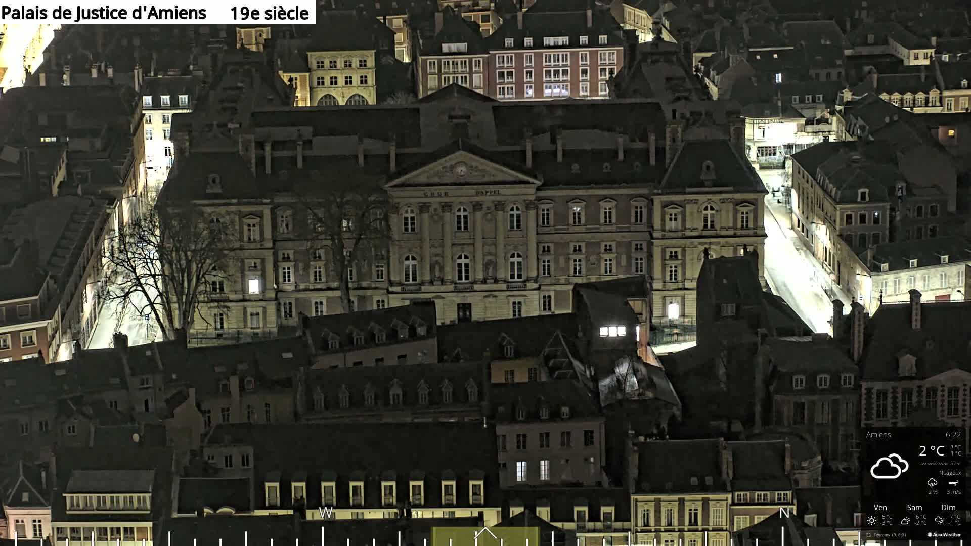 A nighttime aerial view of a European city shows a large, well-lit stone building surrounded by smaller, darker buildings under a cloudy sky.