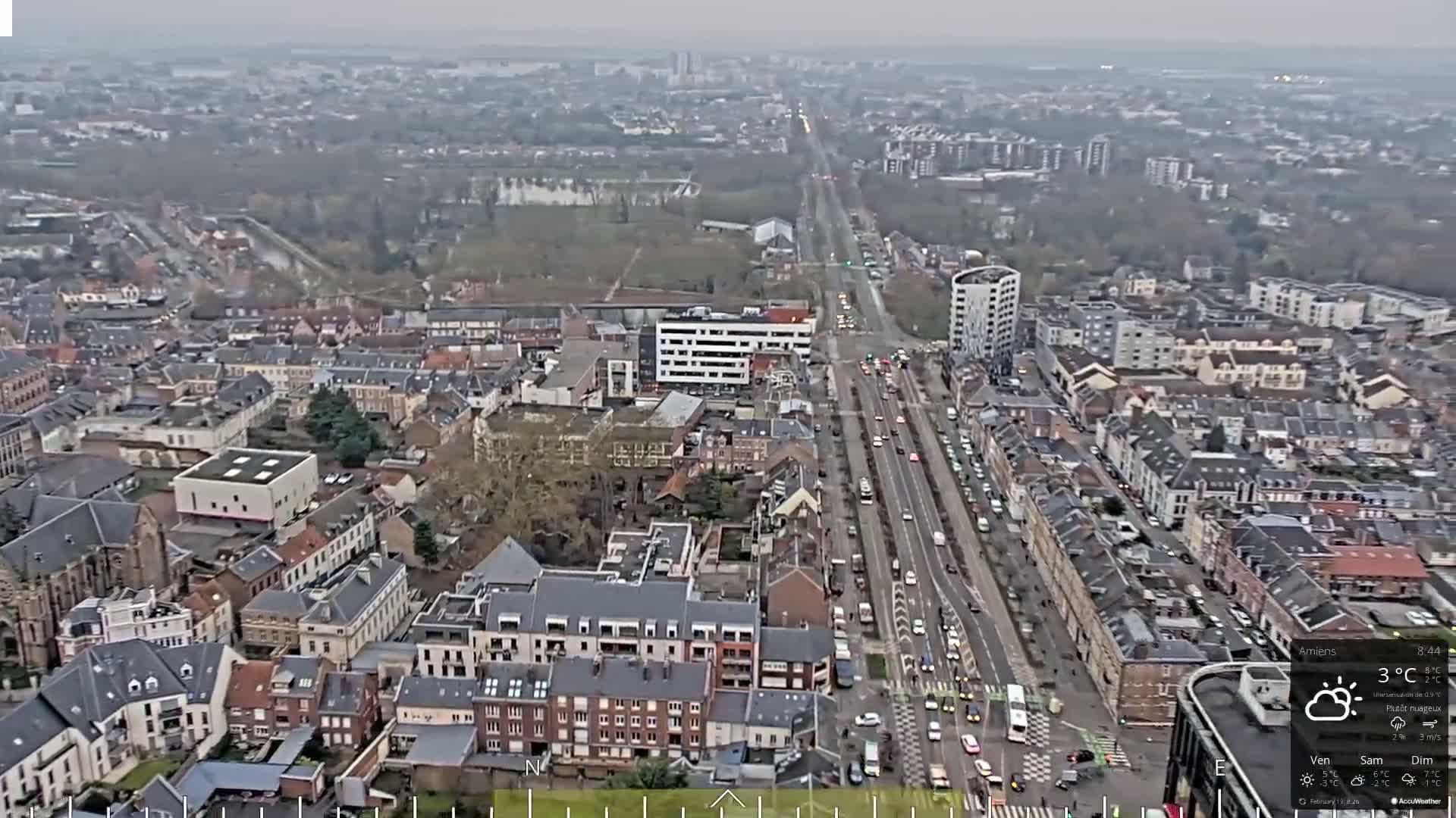 An aerial view shows a city with a long road filled with cars under a mostly cloudy sky.