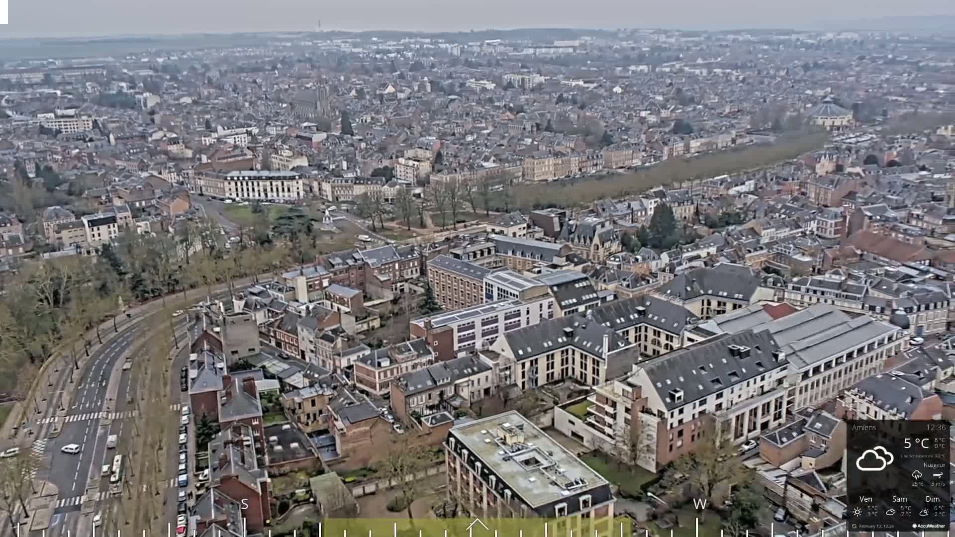 An aerial view of a city on an overcast day shows a sprawling network of streets and buildings, with a road curving through the scene.