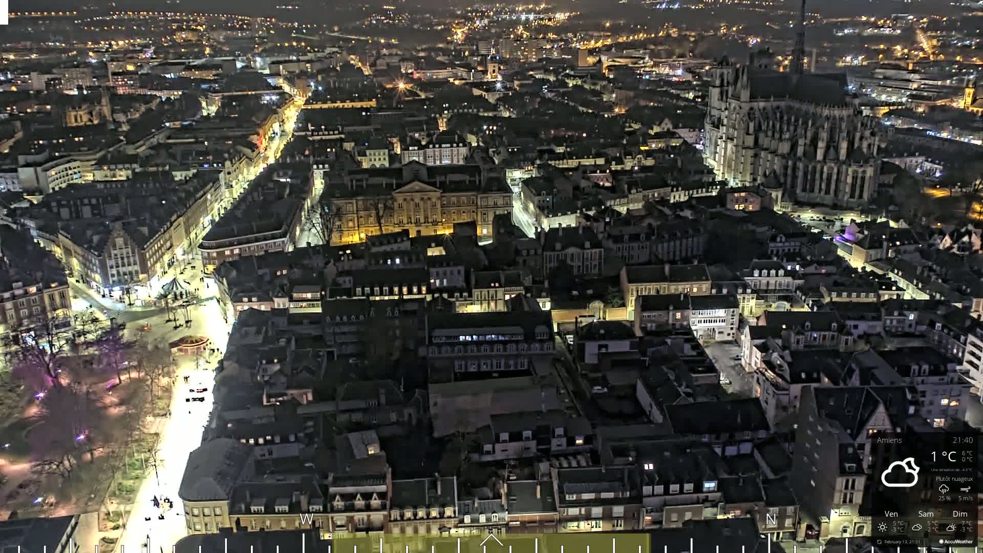 A nighttime aerial view of a city with a large cathedral, illuminated streets, and a mostly cloudy sky.