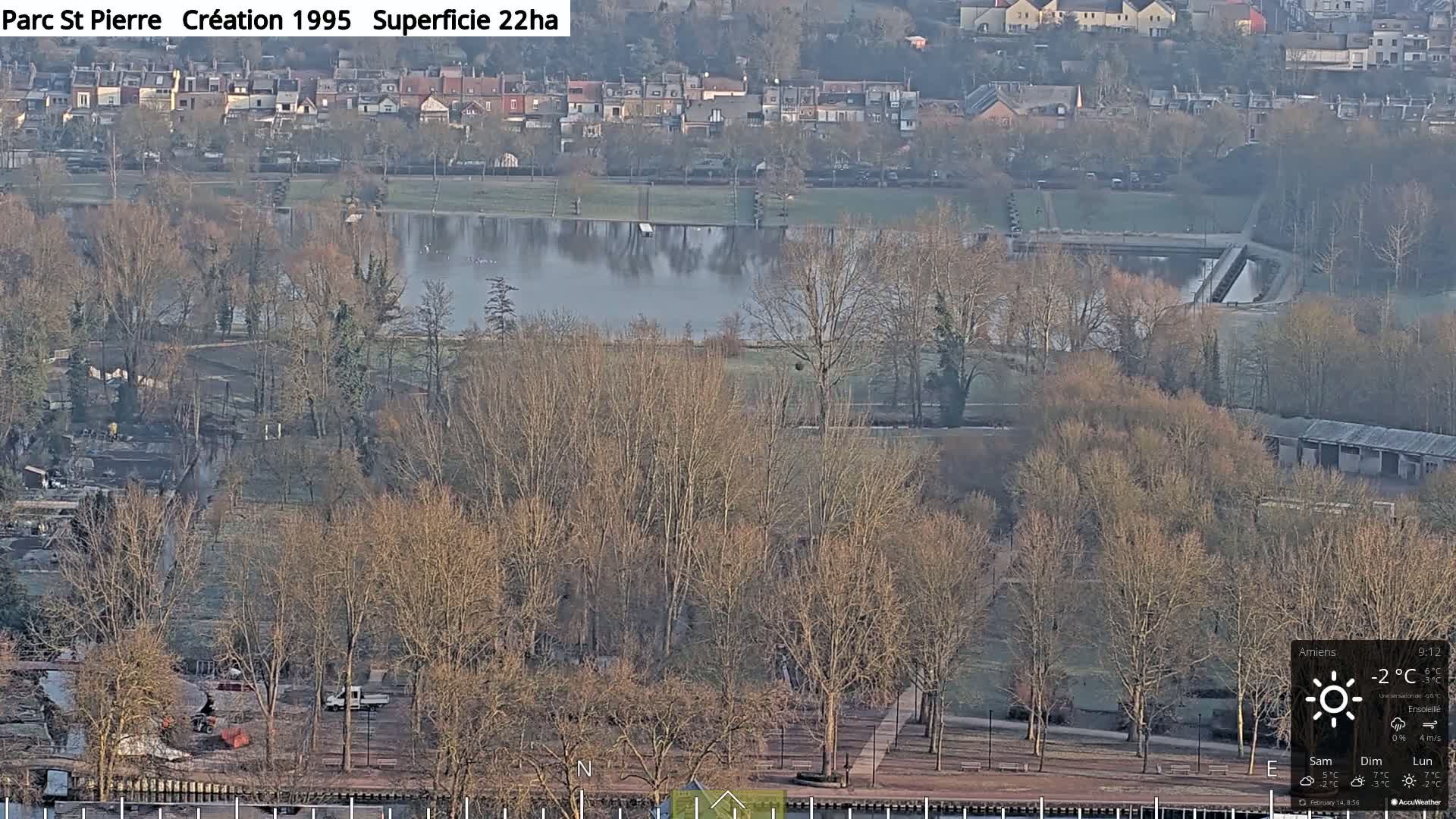 A high-angle, long shot of a park with a lake, bare trees, and a residential area in the background, on a sunny, cold day.