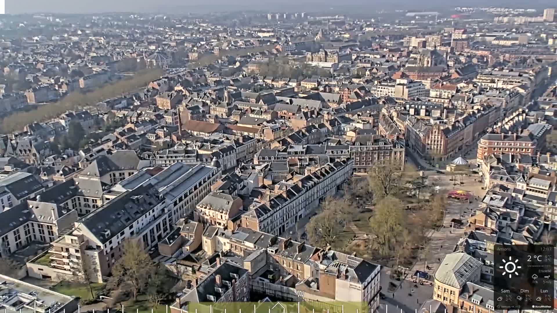 An aerial view of a city on a sunny day shows many closely packed buildings of various sizes and architectural styles, with a large open square or park visible in the center.