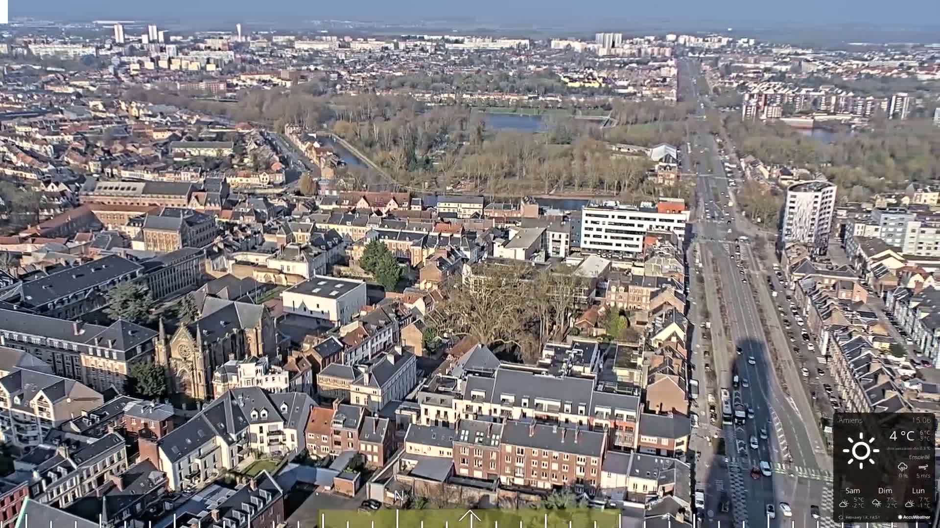 A high-angle, long shot of a European city on a sunny day with a temperature of 4°C, showing numerous buildings, streets, and a large park-like area with a body of water.