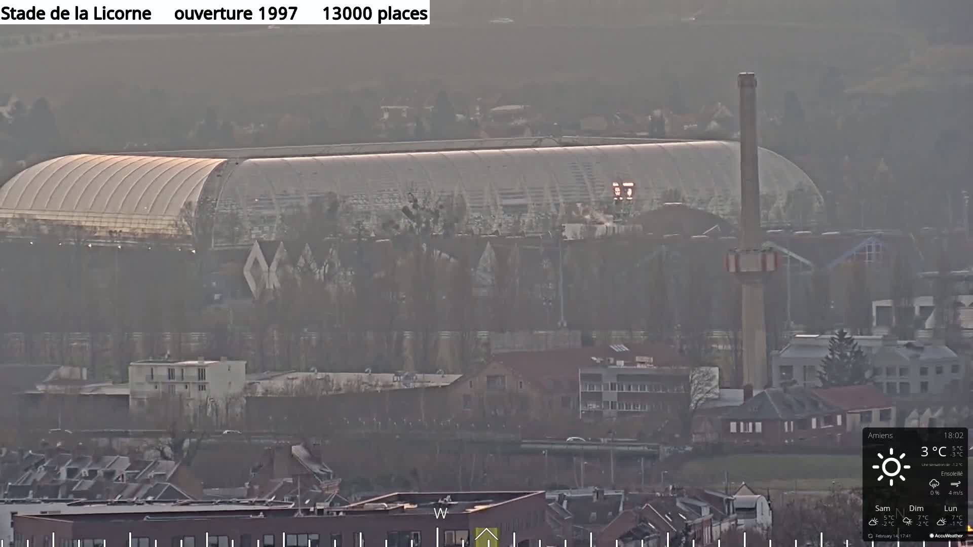 A large stadium with a curved roof is visible in the distance on a hazy, sunny day.