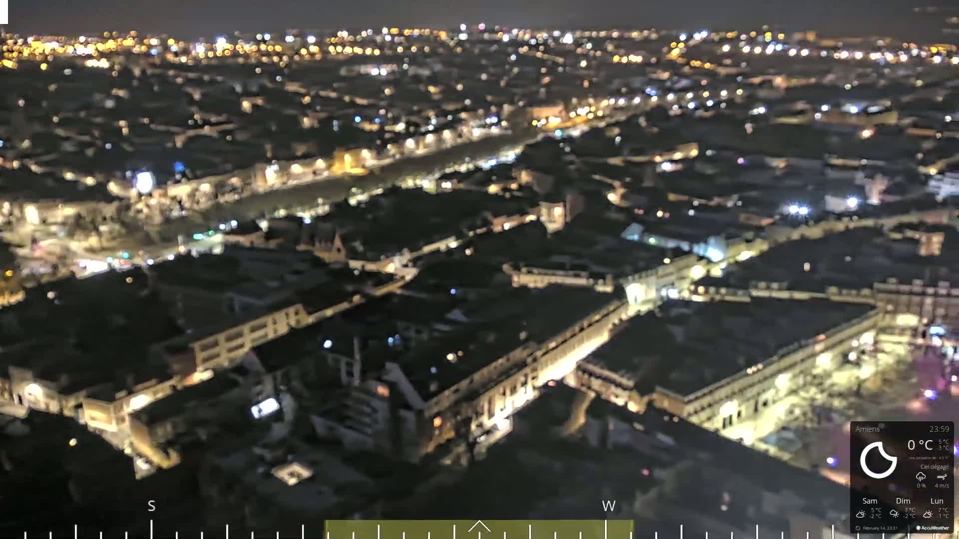 A nighttime aerial view of a city illuminated by numerous lights, with clear skies and a temperature of 0°C.