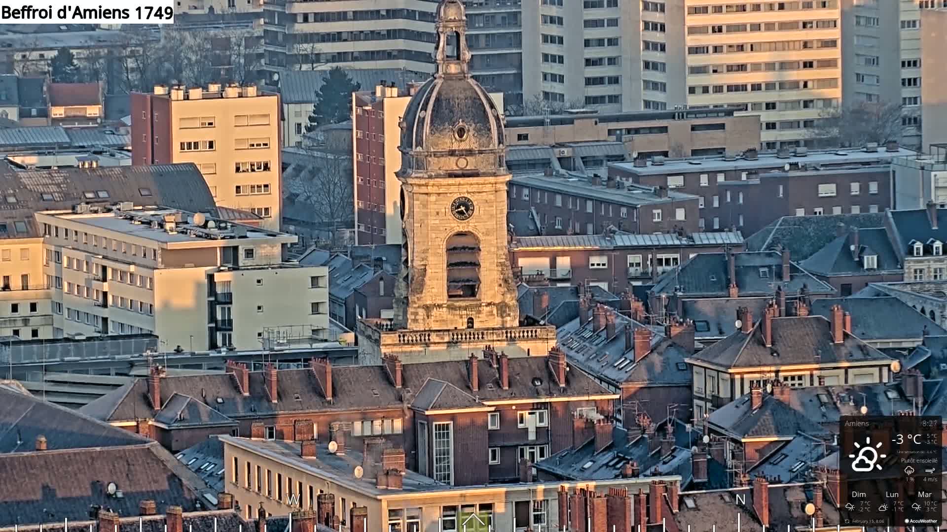 A clock tower stands prominently among a dense collection of rooftops in a city, bathed in the late afternoon sun of a mostly sunny day.