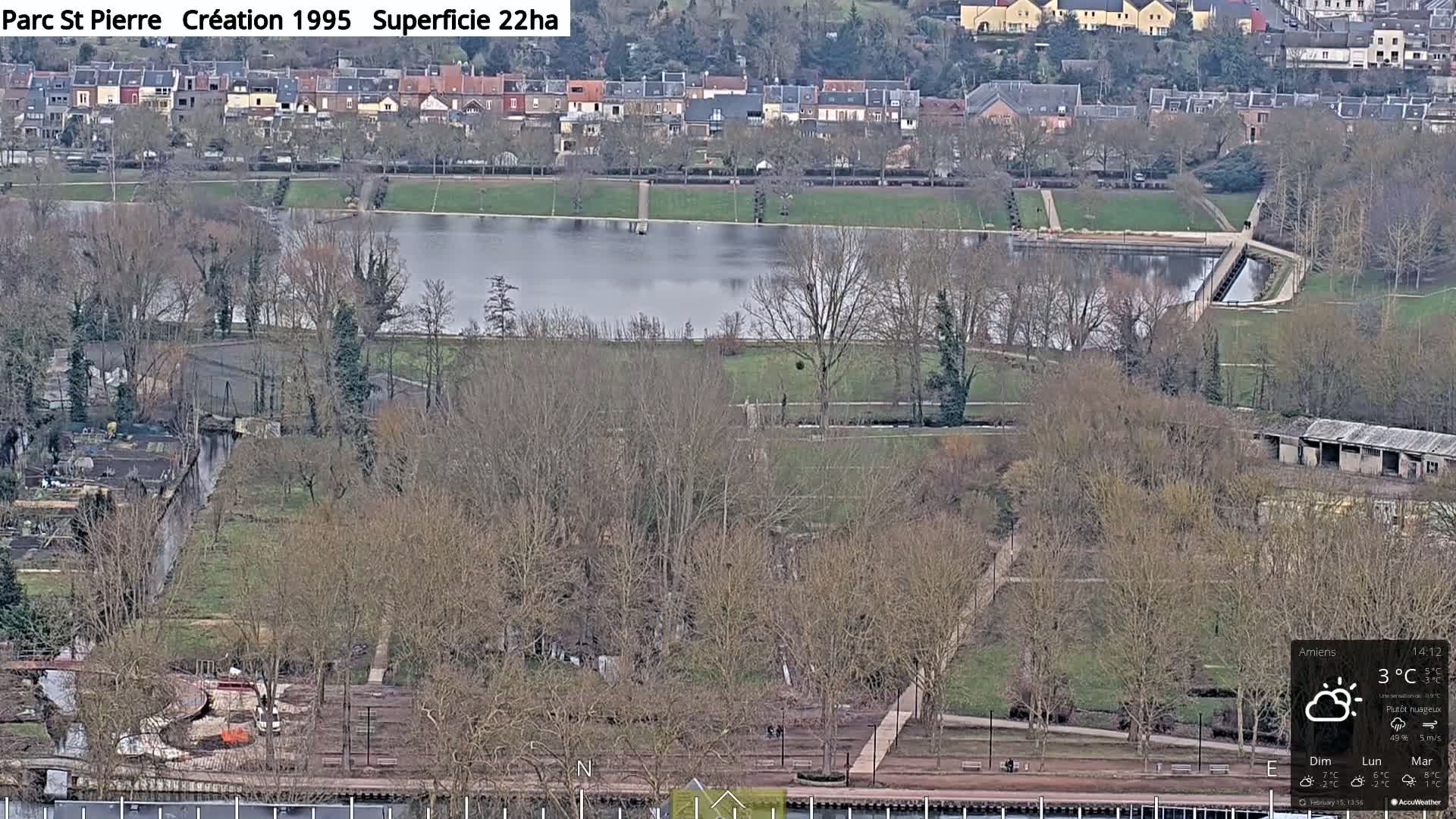 A mostly cloudy day overlooks a park with a lake, walking paths, and bare trees, situated next to a residential area.