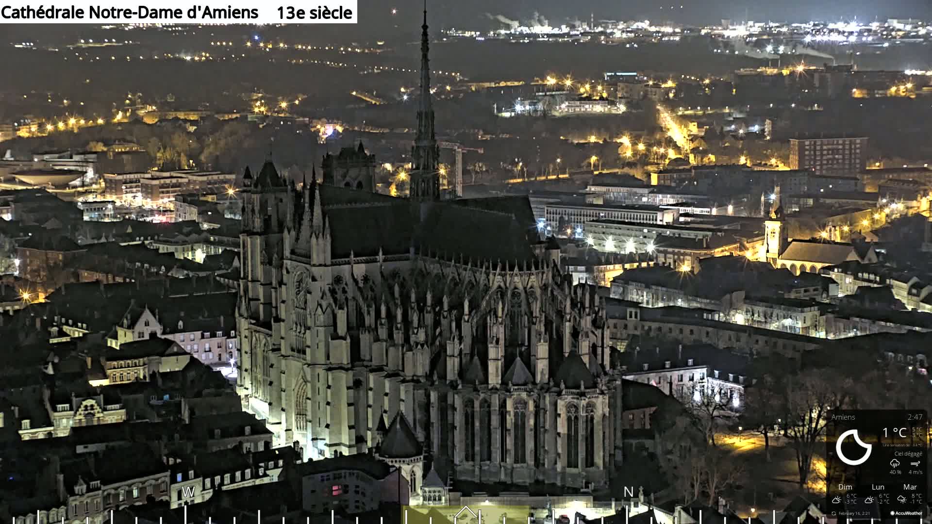 A large Gothic cathedral is illuminated at night, overlooking a city with many lit buildings and streets under a clear sky.